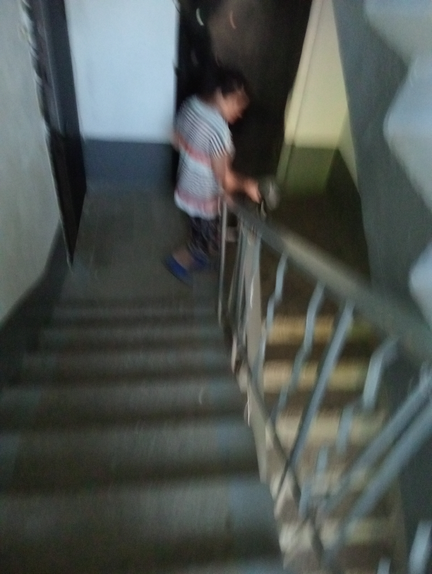The image shows a person on a staircase. The person is wearing a striped shirt and dark pants, with blue shoes. They are holding a dark object, possibly a tool or a container, in their right hand. The staircase appears to be indoors, with a metal railing. The lighting is dim, and the image is somewhat blurry, suggesting it may have been taken in low light conditions or with a moving camera. The scene is enclosed, with walls visible on either side of the staircase. There is no visible text in the image. The activity taking place is unclear due to the blurriness, but the person seems to be either going up or down the stairs while holding an object. The overall impression is that of a candid, possibly utilitarian, shot within an everyday setting.