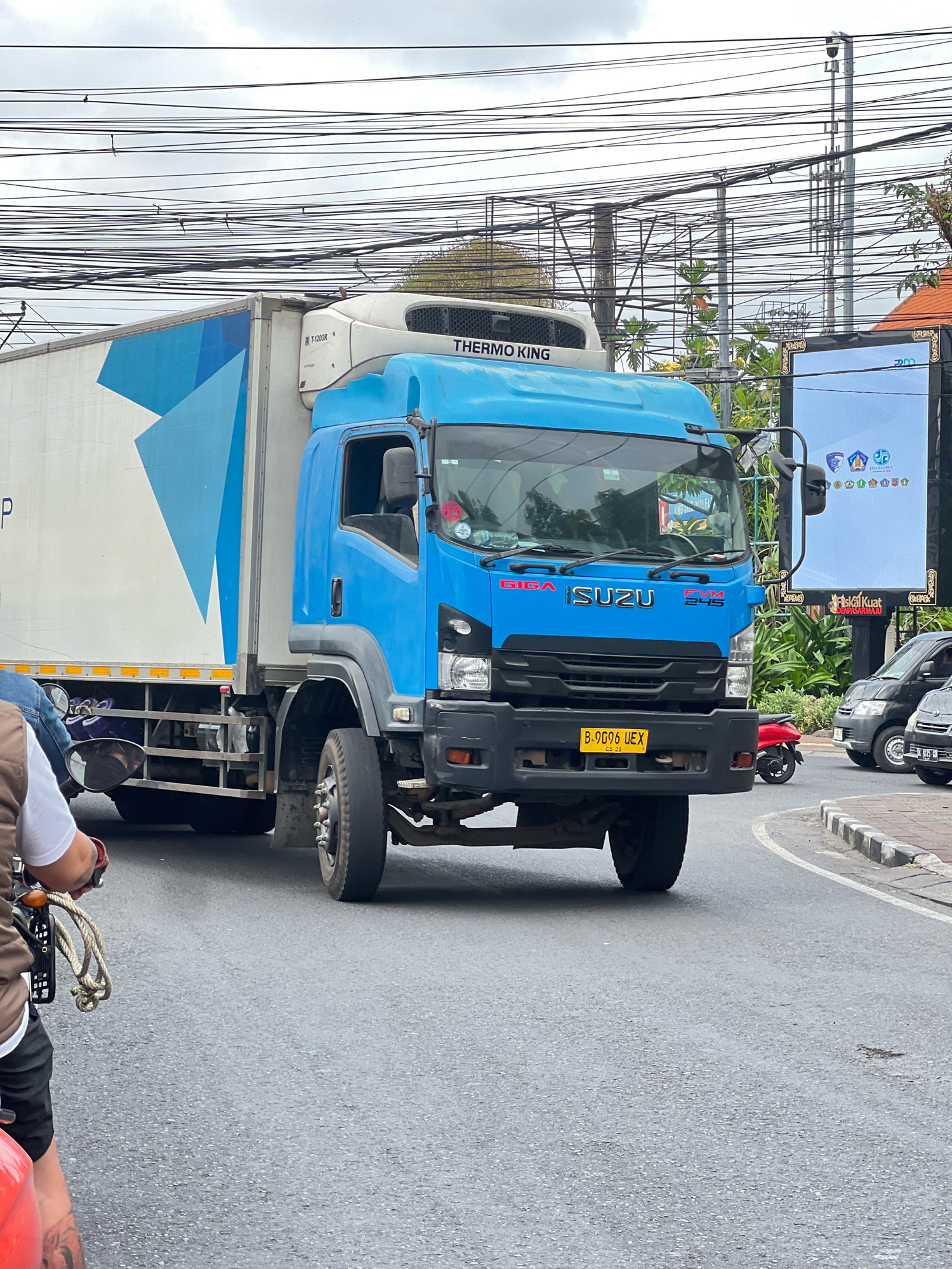In Denpasar, Indonesia, on a cloudy day, a bright blue Isuzu Giga FVM 245 refrigerated truck with "THERMO KING" branding is driving on an asphalt road. The truck has a white cargo container with a geometric blue design on its side. A person on a scooter, wearing a white shirt and brown vest, is in the foreground to the left, holding the scooter's handlebars. Behind the truck, a large digital billboard displays various logos and text, including "ASKAL KUAT" and what appears to be Indonesian text. Other vehicles, including a black van and a red scooter, are visible on the road. Overhead, a complex network of electrical wires crisscrosses the sky. The environment suggests an urban setting with typical city infrastructure.