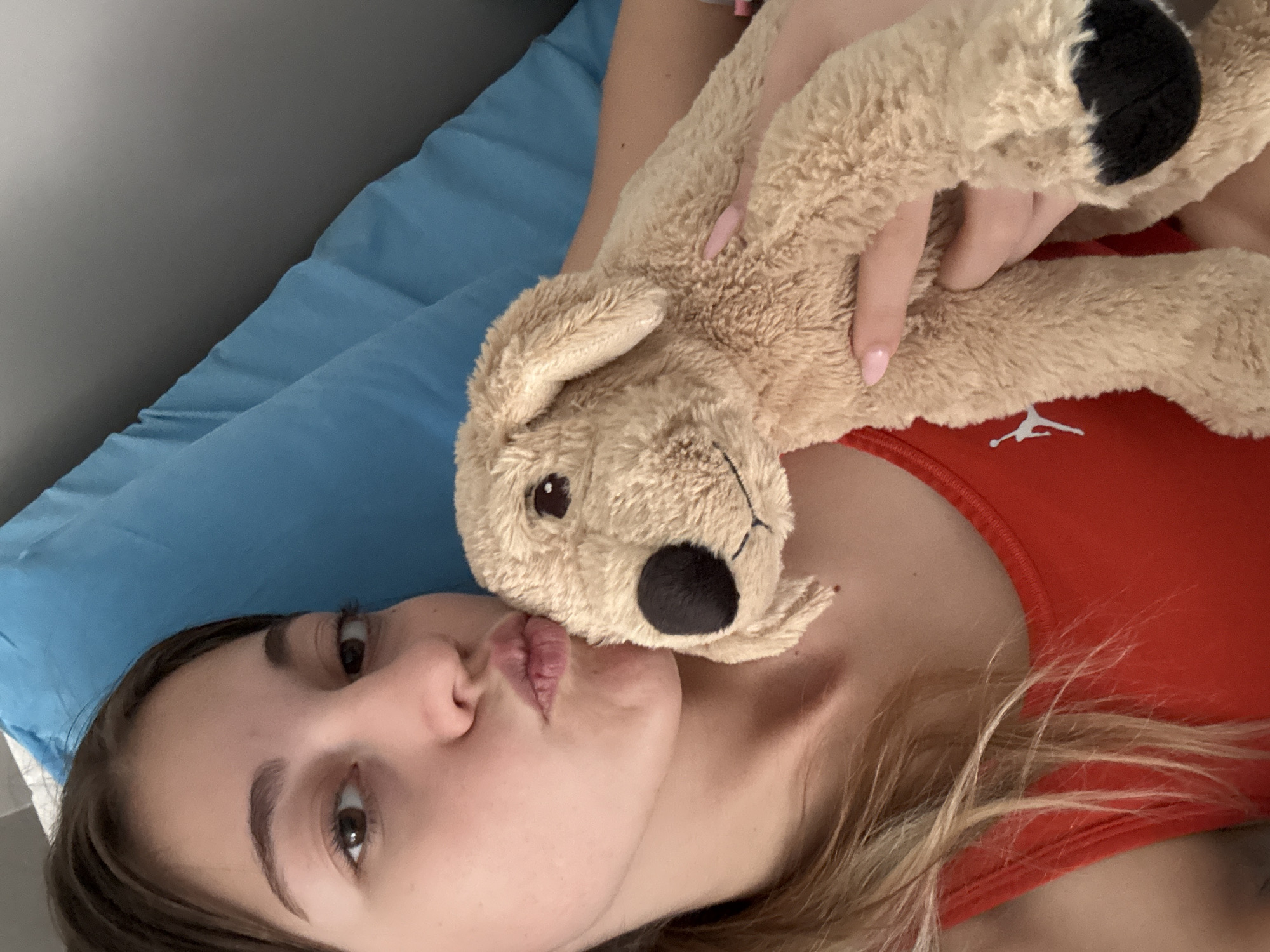 A selfie-style, indoor photograph, likely taken with a mobile device, depicts a young woman lying on her back on a blue pillow. She is wearing a red tank top with a white logo visible on the right side of her chest. The woman is holding a tan, stuffed animal dog against her chest, with her right hand visible holding the dog's paw. Her face is close to the camera, and she is puckering her lips in a kissing gesture. The background is minimally visible, showing a grey or neutral-colored wall. The location in the city of Sofia, Bulgaria cannot be determined from the available image.