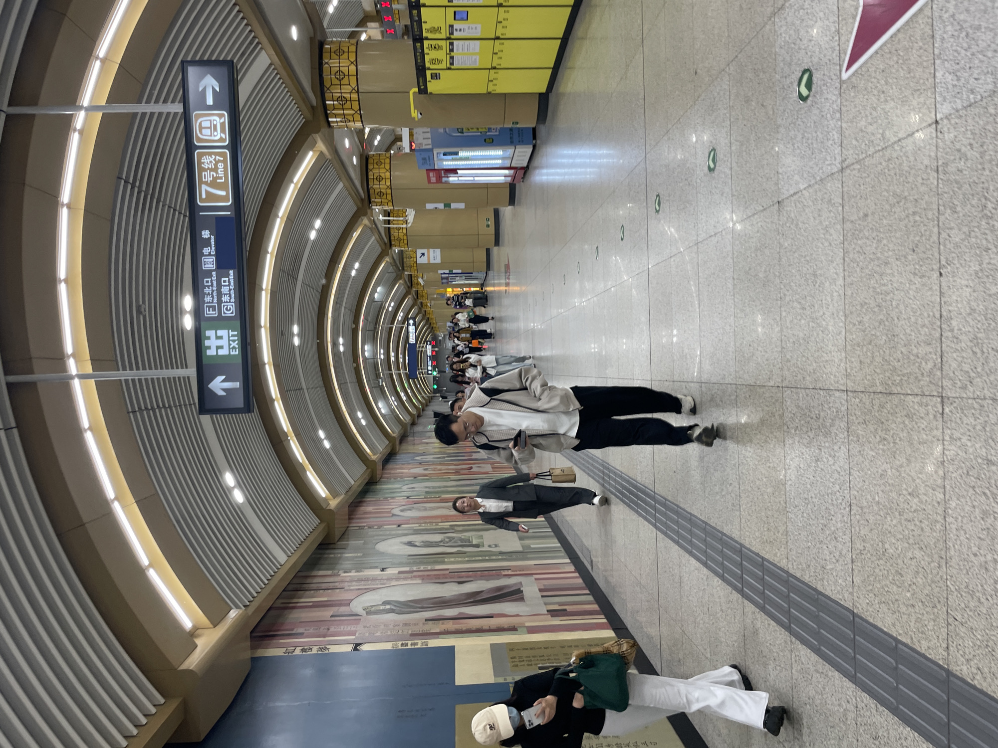 The image shows an underground subway station in Dongcheng District, China, indicated by a sign for Line 7 and exit information in Chinese characters. People are walking along a tiled floor, some looking at their phones. A tactile paving strip runs parallel to the wall, which is adorned with murals or artwork. On one side, there are yellow lockers or vending machines. The station has a curved ceiling with recessed lighting. A sign indicating the location of the elevators is visible, providing guidance to subway users.