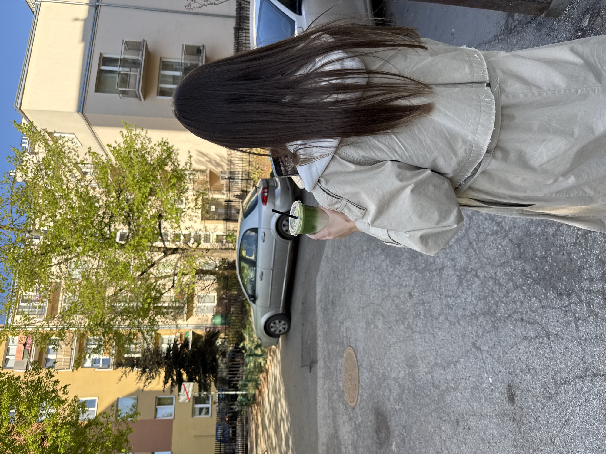 A medium shot shows a person with long, dark hair holding a green drink in a clear plastic cup with a black straw. They are wearing a light-colored jacket. The backdrop includes a multi-story building with light-colored walls and various windows, partially obscured by green trees. A silver car is parked nearby. The scene appears to be a street or sidewalk, with gray asphalt visible. The location is in Warsaw, Poland.