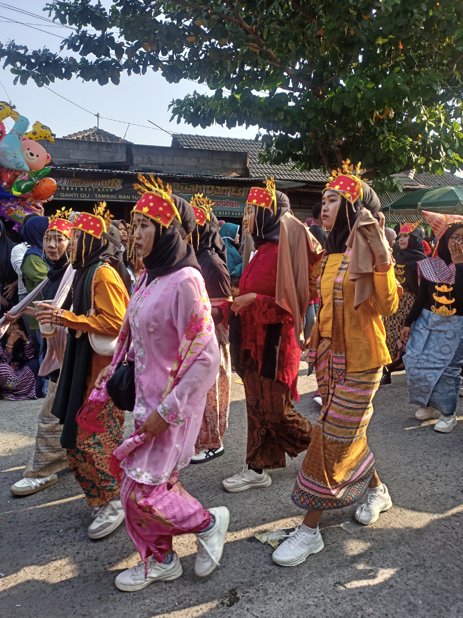 In the city of Saren, Indonesia, a group of women dressed in traditional attire participates in a parade. The women, some of whom are wearing vibrant red and gold headpieces adorned with intricate gold detailing, are seen walking on a paved street under a clear sky, partially shaded by lush green trees. Their clothing consists of brightly colored fabrics, including a pink dress with floral patterns, an orange tunic, and patterned skirts. Some women are also wearing black hijabs. In the background, a building with a tiled roof and a sign that reads "DIMOTOR" is visible, along with a vendor selling a cluster of colorful balloons. The women are walking from left to right in the frame, with some engaging in conversation or holding drinks. The atmosphere appears festive, suggesting a cultural celebration or event.