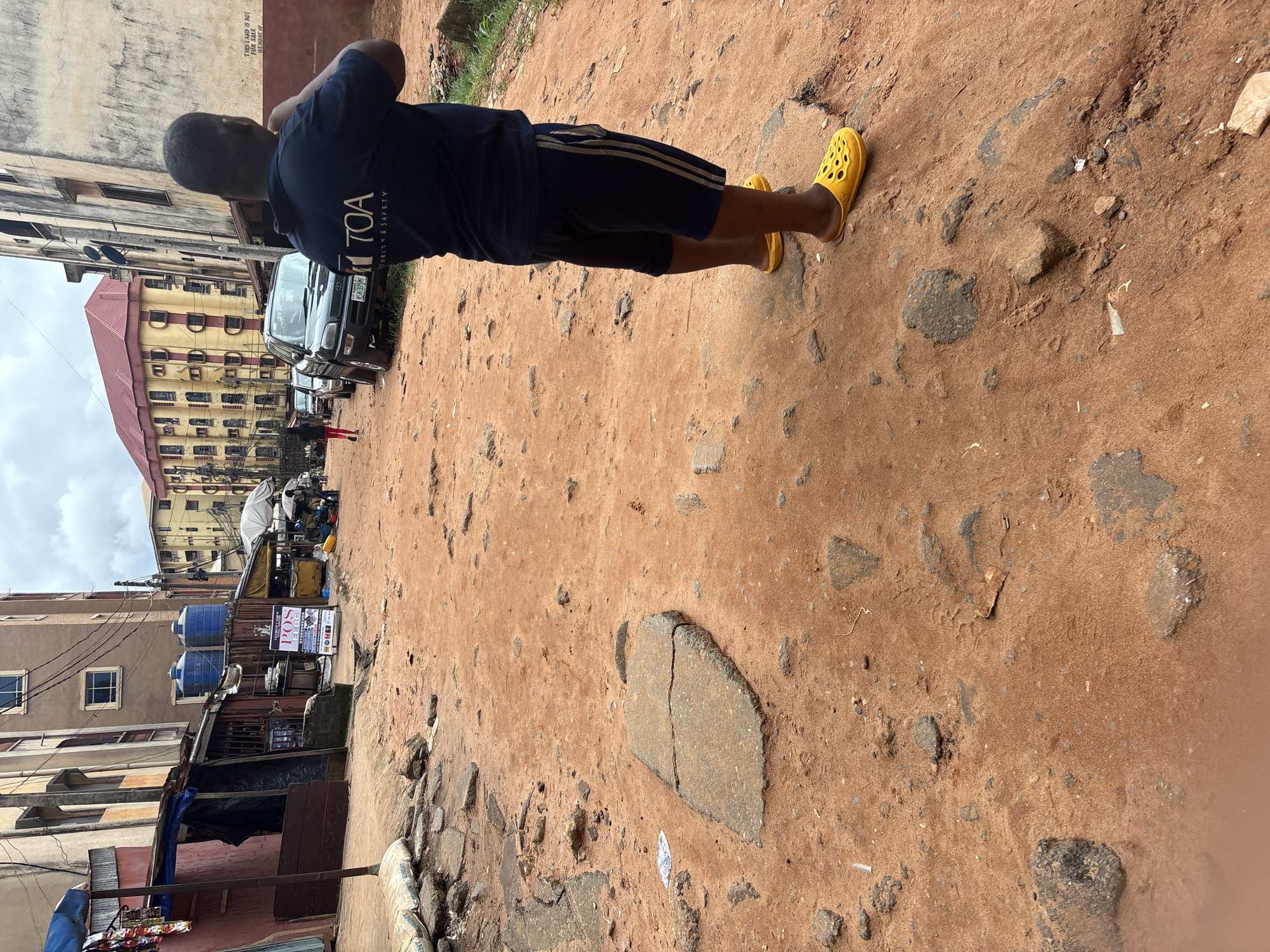 The image captures a street scene in Onitsha, Nigeria, characterized by a red-dirt road and surrounding buildings. In the foreground, a man is viewed from behind, wearing a dark blue t-shirt with the "TOA SECURITY & SAFETY" logo and dark blue shorts with lighter stripes. He is also wearing yellow clogs. The road is uneven and appears to be under construction or in disrepair, with scattered stones and debris.
To the left, there are structures that appear to be market stalls or shops, some with blue tarpaulin coverings. One stall features a sign with "POS" written on it, indicating a point-of-sale service. Above these stalls, large blue water tanks are visible on a building.
In the mid-ground, a black car is parked on the side of the road, and further down the street, other vehicles and a person in a red shirt can be seen. The background is dominated by multi-story buildings with yellow and beige facades, and a red-tiled roof on a prominent building. The sky is overcast. A white sign on a building on the right side of the image reads "THIS LAND IS NOT FOR SALE - BEWARE OF...". The overall impression is of a busy, potentially commercial area in an urban setting with visible infrastructure challenges.