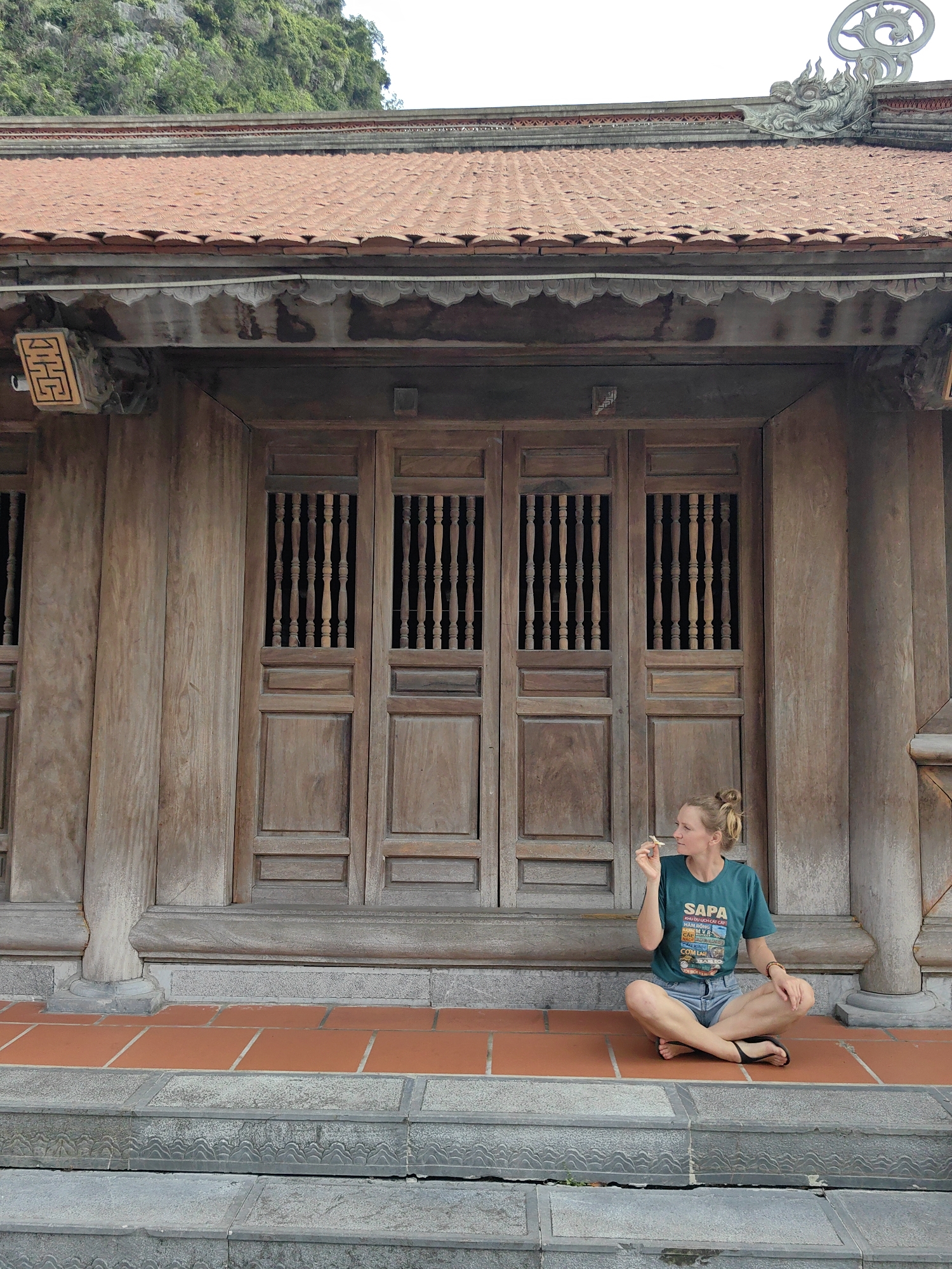 A young woman sits cross-legged on the terracotta tiled porch of a traditional Vietnamese building in Hong Gai Ward, Vietnam. She is dressed casually in a dark teal t-shirt, light denim shorts, and black flip-flops. Her blonde hair is tied in a bun, and she wears a beaded bracelet on her left wrist. She holds a small, light-colored object in her right hand and gazes thoughtfully to her right.
Behind her, the building showcases intricate wooden architecture, including large double doors with ornate wooden balusters in their upper panels. The roof is distinctive with its reddish-brown scalloped tiles and grey stone decorative elements, notably a dragon head sculpture and a circular finial at the peak. Ornate carvings adorn the underside of the eaves, beneath which a modern white wire is visible. The building rests on a foundation of grey stone, with terracotta floor tiles leading to a set of grey stone steps, which feature a delicate wave-like carved pattern. In the background, a verdant, rocky mountain rises under what appears to be an overcast sky, indicating daytime.
Her dark teal t-shirt prominently displays "SAPA" along with "KHU DU LICH CẮT CÁT" and other smaller Vietnamese text, accompanied by illustrations of various vehicles. The scene suggests a tranquil, contemplative atmosphere.