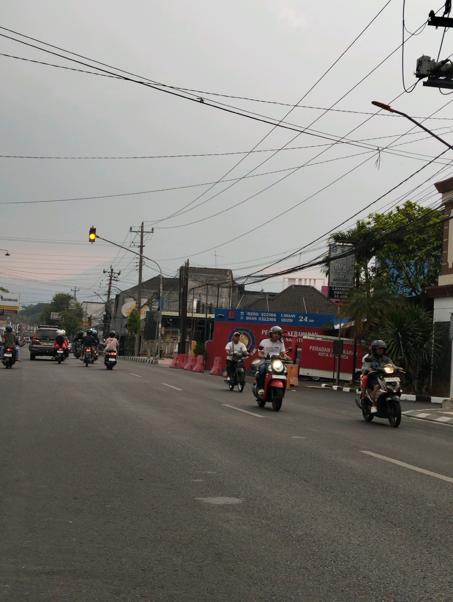 The image captures a street scene in Salatiga, Indonesia, with a traffic light showing yellow. Several motorcycles and a car are visible on the asphalt road. In the background, buildings and trees line the street, and a dense network of power lines crisscrosses the overcast sky. Two men on a red scooter are in the foreground, with another person on a black and white scooter to their right, also on the road. A man is riding a motorcycle in the background, and behind him, a silver car is driving. Further down the road, several other motorcycles are seen. A street sign is visible on the right side of the street.