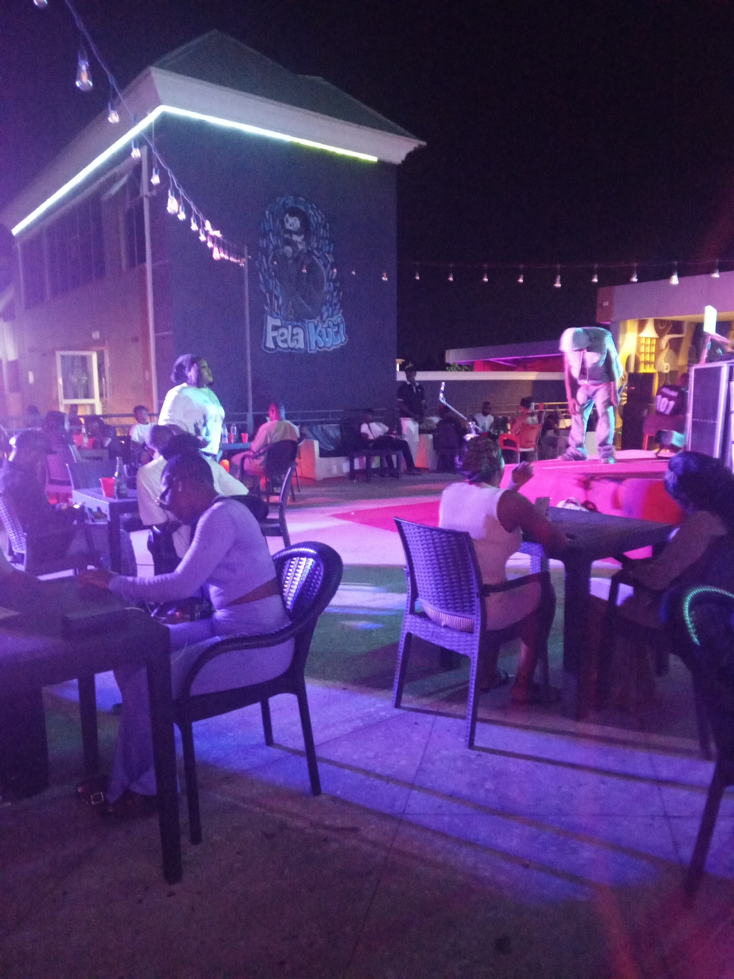 Here is a detailed description of the image:
**Scene:** An outdoor nighttime scene, likely a bar or restaurant with a stage area. The location is indicated by a mural of Fela Kuti, suggesting it might be in Lagos, Nigeria or a place celebrating his music.
**People:** Several individuals are present, mostly seated at tables.
* A woman seated at a table is looking down, possibly at her phone. She is wearing a long-sleeved top and pants.
* Another woman stands behind her.
* A woman in white sits with her leg raised.
* On the stage, there appears to be a person bending over. Another person with "101" on their shirt is standing near speakers.
* Other patrons are seated at various tables in the background.
**Objects:**
* Tables and chairs
* String lights overhead, providing illumination.
* A stage with speakers and other equipment.
* A large mural of Fela Kuti on a building.
* Drinks on tables.
**Actions and Interactions:**
* People are seated and appear to be talking or watching the stage.
* The individual on stage is bending over, possibly adjusting equipment or performing.
**Environment:**
* The scene is outdoors with a tiled or paved surface.
* The lighting is low, with purplish and blue hues, likely from stage lighting or decorative lights.
* The atmosphere seems lively with people enjoying the evening.