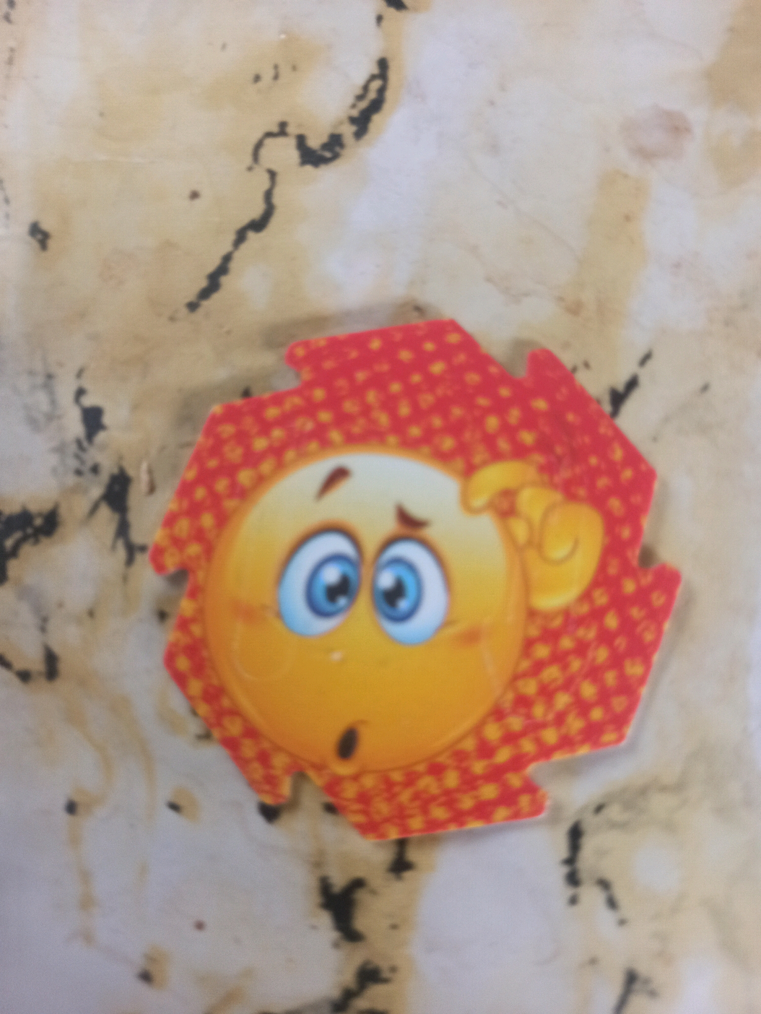 A hexagonal, cartoon-like image of a yellow emoji with blue eyes and a shocked expression is placed on a light-colored, textured surface with dark smudges. The emoji has a red and orange patterned border. The scene is indoors, and there is no discernible connection to the city of Machraa El Ain, Morocco.