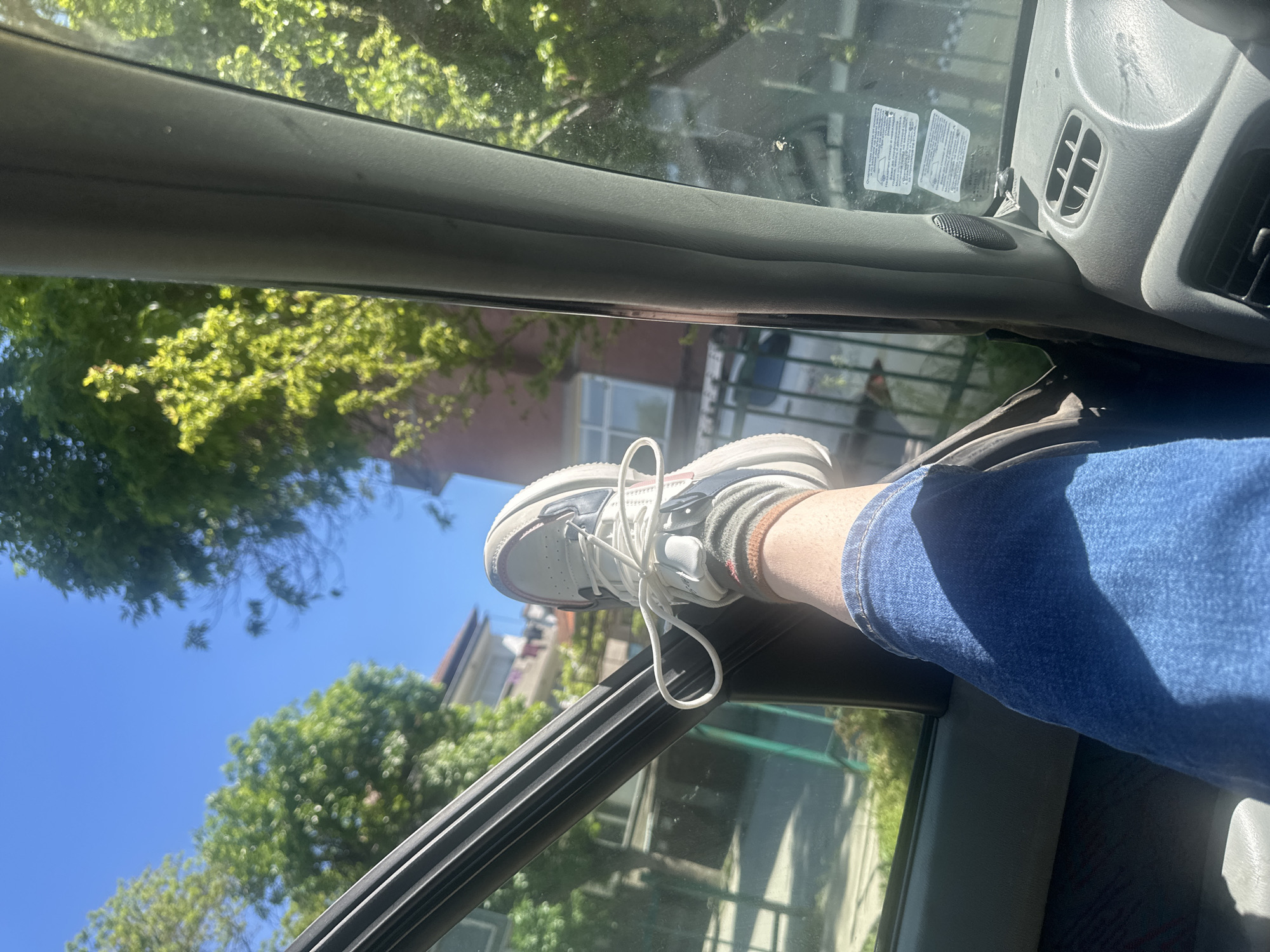 A first-person view looking out from inside a vehicle. A leg wearing blue jeans and a sock-clad foot wearing a white athletic shoe is propped up on the car door. Exterior residential buildings and deciduous trees are visible through the car window. The scene appears to be outside, with clear blue sky above and ample natural light. The location cannot be identified.