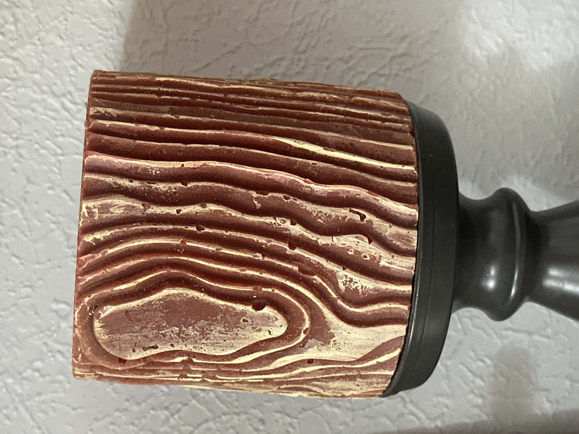 The image shows a close-up of a candle holder with a candle on top. The candle is cylindrical and has a textured surface that mimics the grain of wood. It is red-brown with lighter, yellowish-beige highlights in the grooves. The candle rests on a black, pedestal-style candle holder. The background is a textured, light-colored wall. The scene is indoors, and there is no indication of the city of Nevinnomyssk, Russia.
