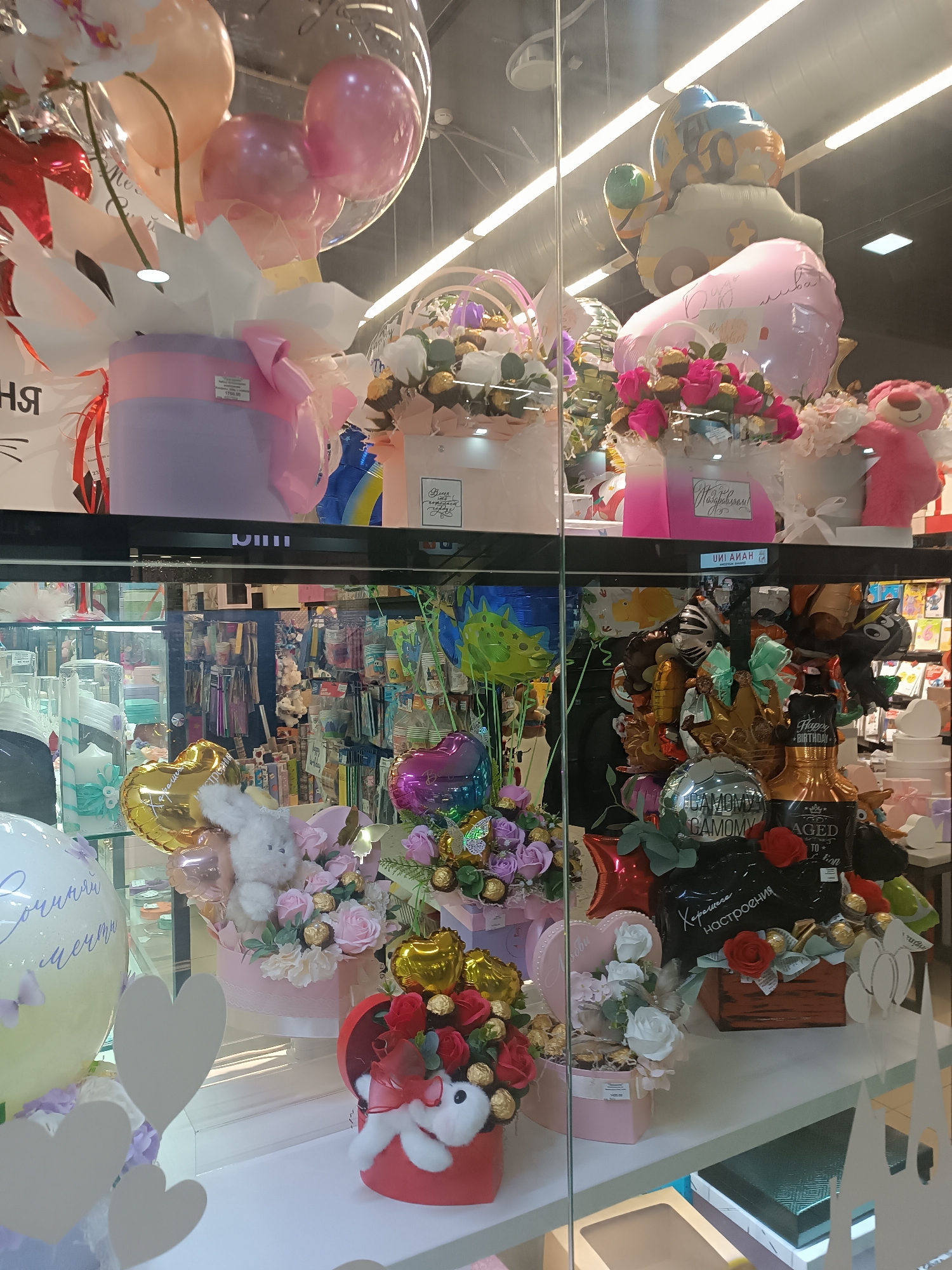 Here's a detailed description of the image:
**Scene:** The image depicts a display of gift arrangements in what appears to be a shop window. The items are situated on two shelves behind a glass barrier. The location is identified through Cyrillic text in the image which translates to "Sepych," placing it in the city of Sepych, Russia.
**Objects:**
* **Top Shelf:** Displays arrangements consisting of balloons, gift boxes, roses, chocolates and stuffed animals.
* **Bottom Shelf:** Similar arrangements to the top shelf, including roses, chocolates, stuffed animals, and balloons. Some arrangements include chocolate-covered strawberries or other treats.
* **Balloons:** A variety of balloons including heart-shaped, spherical, and character balloons.
* **Gift Boxes:** Boxes in different shapes (square, round, heart-shaped) and colors (red, pink, lilac, beige).
* **Signs:** There are signs or tags, some with Cyrillic text, including what looks like a happy birthday message and a brand name.
* **Interior View:** Through the glass, the shop's interior is visible, showing more products on shelves, including balloons, candles, cups, and gift boxes.
**Environment:**
* The shop display is well-lit.
* The inside of the shop is orderly and stocked with merchandise.
* The foreground view suggests the display is in a retail setting.
**Overall Impression:** The scene is a commercial display of gift arrangements designed for celebrations and special occasions, likely for sale in a shop within the city of Sepych, Russia.