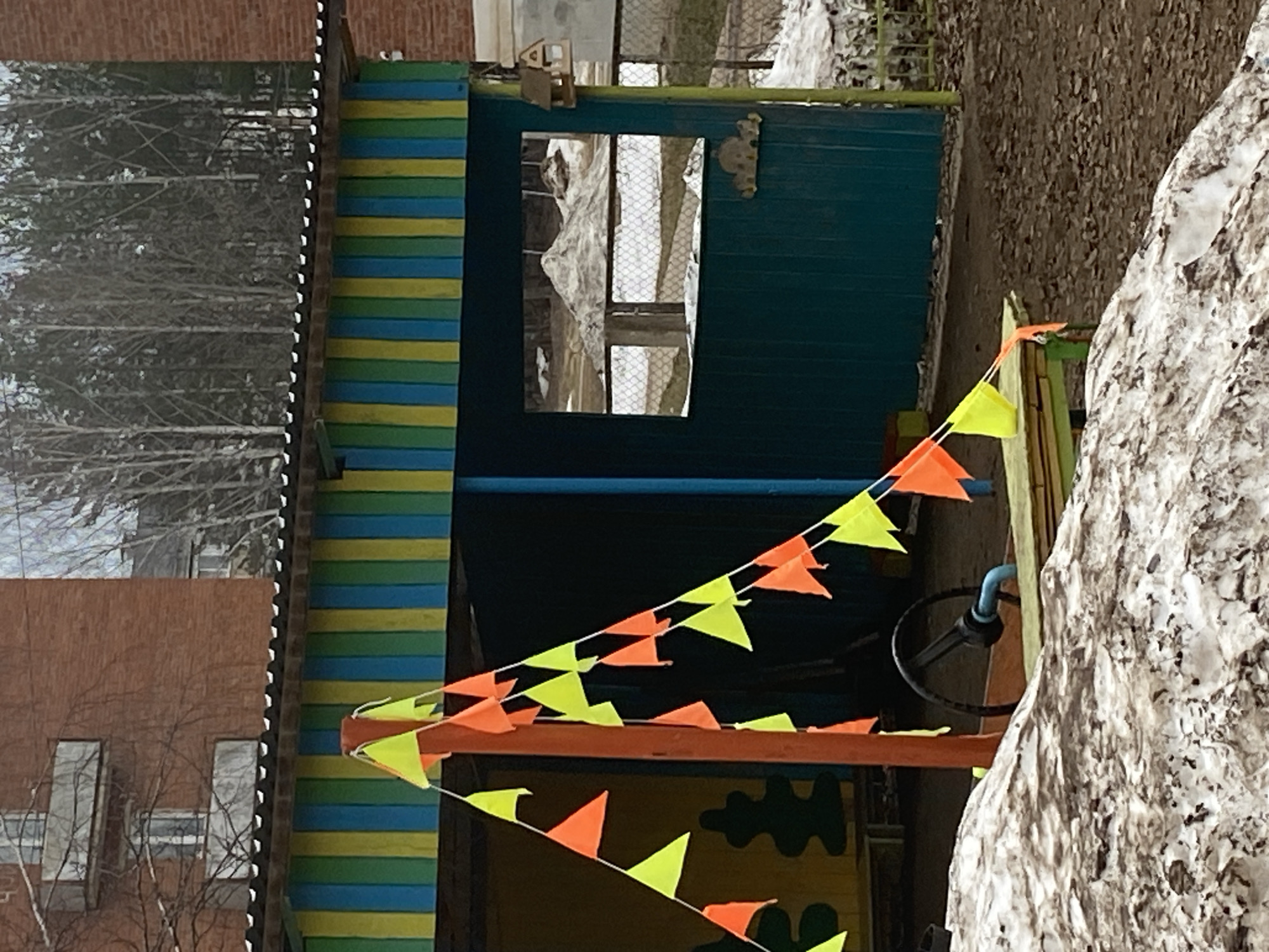 The image depicts an outdoor scene, likely a playground or recreational area, located in Noviy, Russia. A structure painted with horizontal blue and yellow stripes stands adjacent to a brick building. The structure is decorated with a string of alternating yellow and orange triangular pennants. A small, screened window is visible in the structure's wall. In the background, bare trees are visible. A large, textured, possibly snow-covered, rock or hill is present in the foreground, partially obscuring the right side of the structure. An apparent steering wheel can be seen behind the pennants.