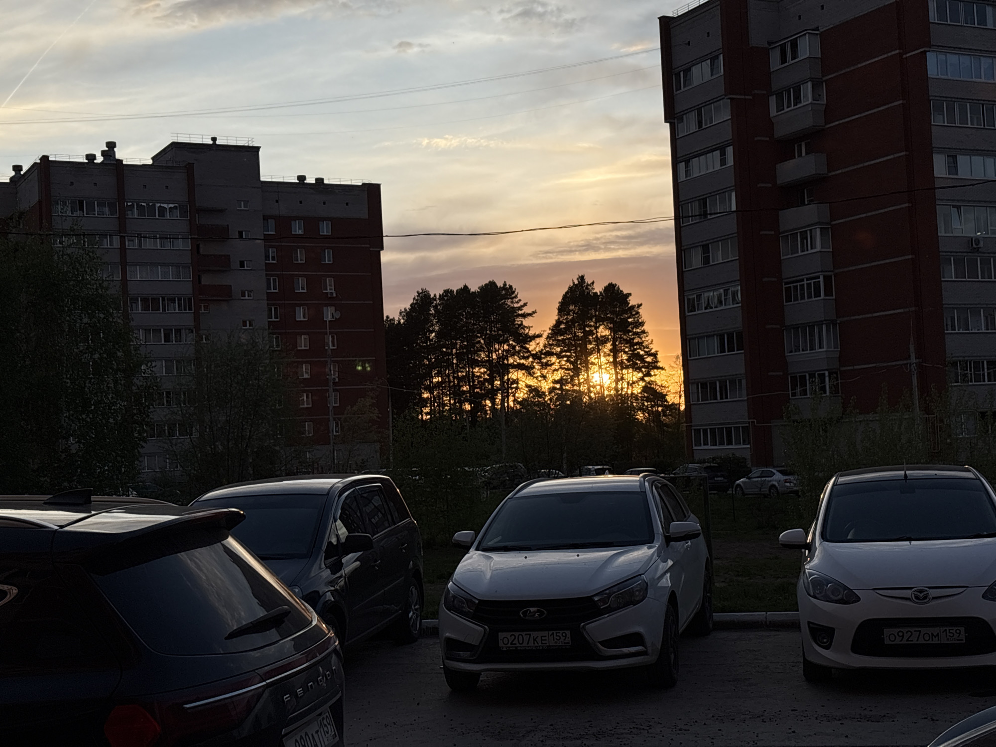 The image depicts a parking lot in Chaikovsky, Russia, at sunset. Several cars are parked, including a black crossover SUV, a dark minivan, a white Lada Vesta with license plate 0207KE159, and a white Mazda Demio with license plate 0927OM159. Behind the cars are trees silhouetted against the setting sun, and in the background are tall apartment buildings. The sky is filled with golden clouds.