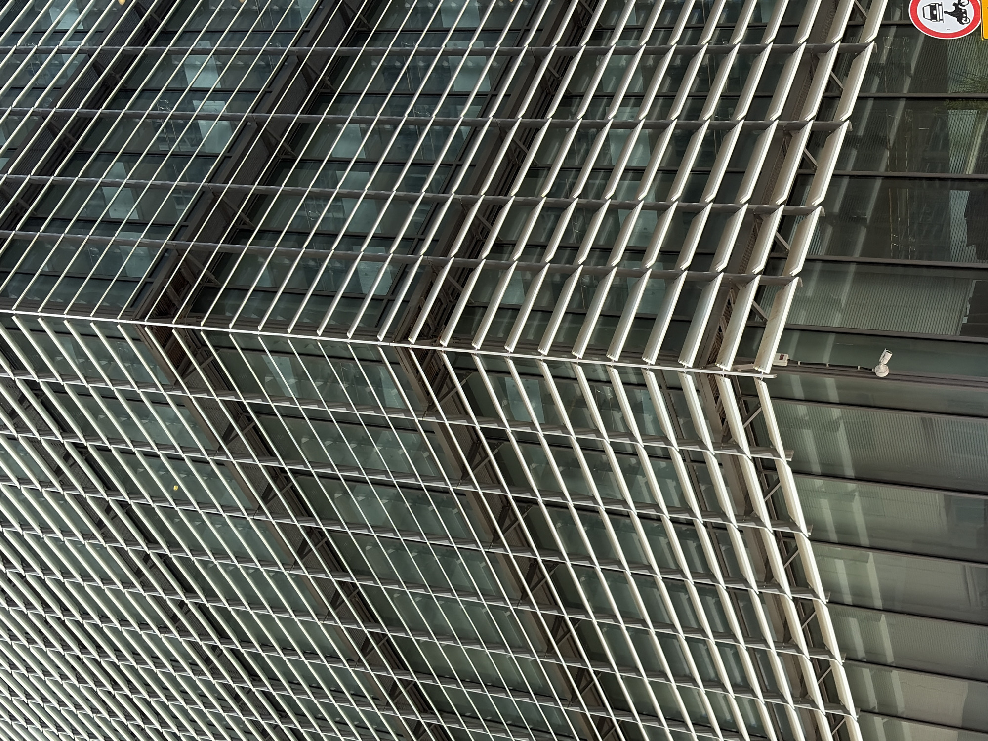 The image depicts an architectural detail of a modern building, likely located in Tel Aviv, Israel. The facade features a grid-like structure of parallel, white metal slats, creating a geometric pattern overlaid onto the glass windows of the building. A security camera is visible, mounted on the wall. There is a sign that prohibits motorcycles. The overall impression is one of contemporary urban design.