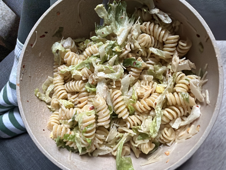 A high-angle, close-up photograph captures a light-colored bowl filled with pasta salad. The salad is composed of spiraled rotini pasta, shredded green lettuce, and a creamy, light-beige dressing. Small red flakes, consistent with chili flakes, are dispersed among the ingredients and adhere to the bowl's interior. The bowl itself appears to be made of a natural-colored material. In the blurred background, a person's lower legs are visible; one leg is covered by a garment featuring green and white horizontal stripes, and the other by dark grey fabric. This setup suggests a casual, indoor environment, likely a private residence, located in New York City, United States of America. No specific city landmarks are discernible within the image.
