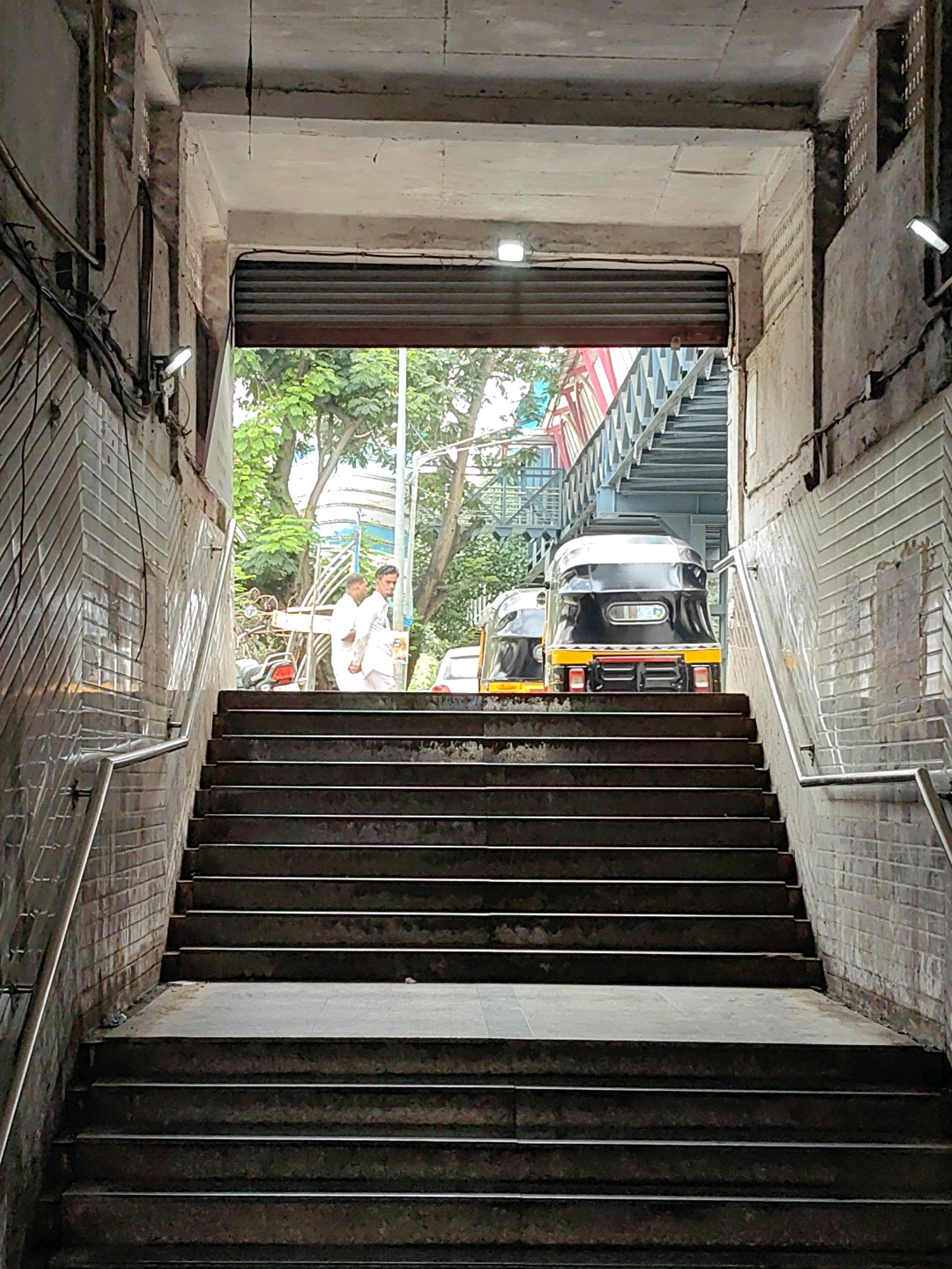A low-angle shot captures a concrete pedestrian underpass with a wide staircase leading upwards. The walls of the underpass are clad in white rectangular tiles with a subtle sheen, and the ceiling is exposed concrete. A metal roller shutter is visible at the top of the underpass, partially obscuring the view beyond. Two figures in white traditional Indian attire are standing on the upper part of the stairs, facing away from the camera. Behind them, a black and yellow auto-rickshaw is parked, with another partially visible to its left. Trees and foliage are visible in the background, suggesting an outdoor environment. The overall scene depicts a busy urban intersection with pedestrian and vehicular traffic.