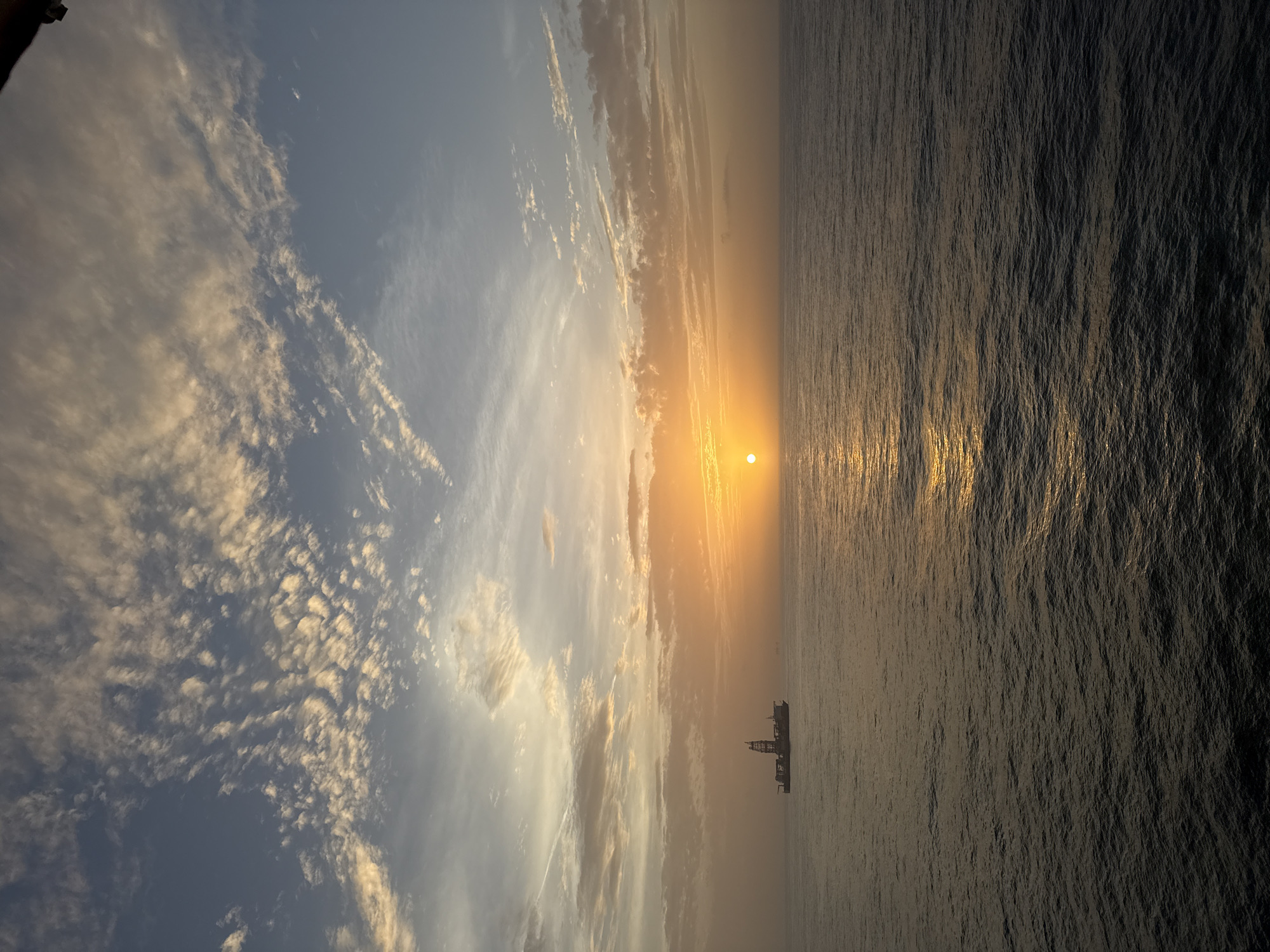 The image depicts a seascape at sunset. The sun is positioned on the horizon, between the sky and the sea, creating a golden glare across the water. The sky is filled with clouds, some reflecting the golden light of the sun. An offshore platform or ship is visible in the distance. The location is Unknown, Unknown.