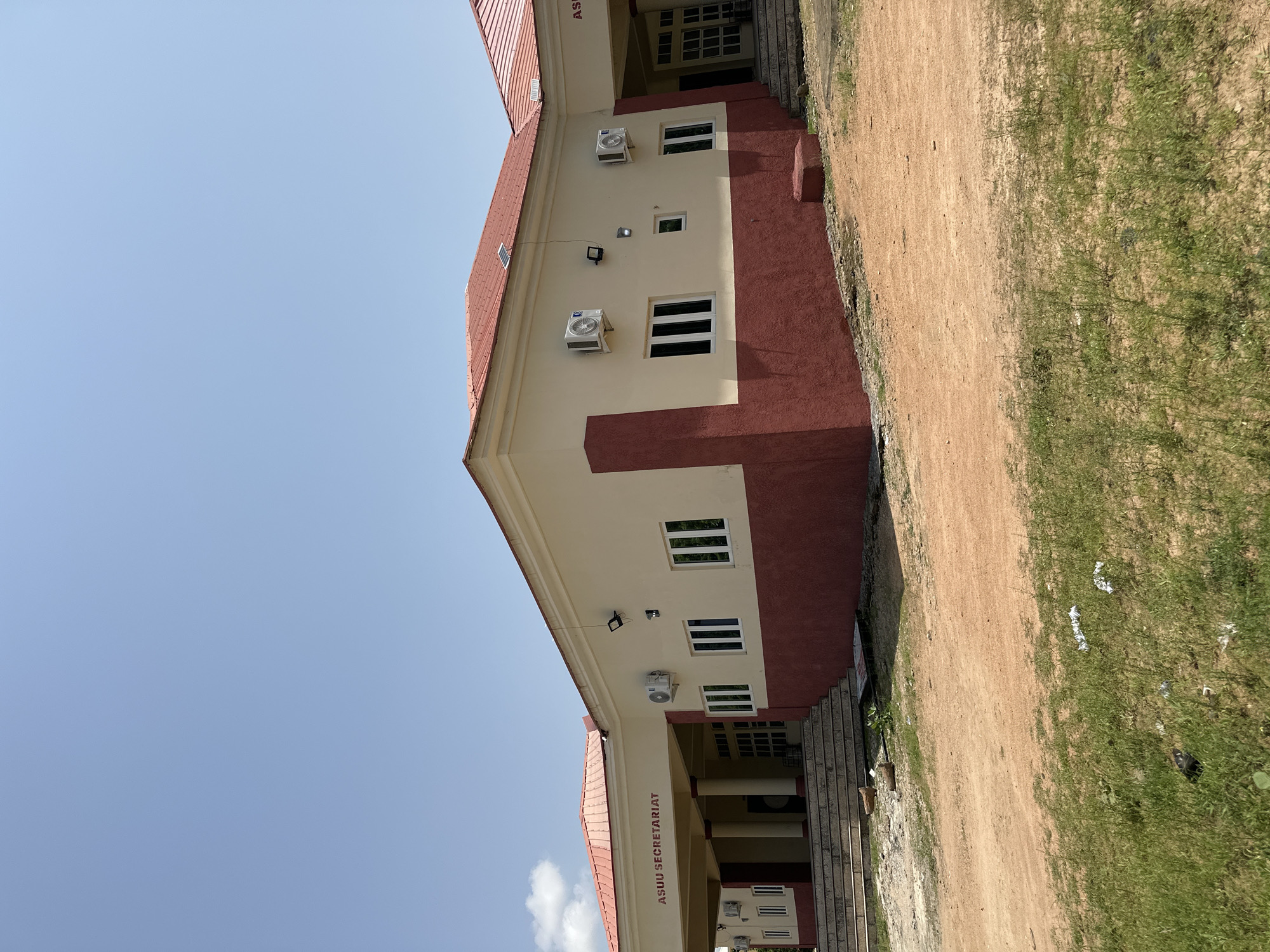 The image shows the exterior of the ASUU Secretariat building in Jalingo, Nigeria. The building has a light yellow facade with a dark red lower section and a red metal roof. Several windows and air conditioning units are visible on the exterior walls. A covered entrance with pillars is also present, with "ASUU SECRETARIAT" written above it. The building is situated on a dirt and grassy area, with some debris scattered on the ground. The sky is clear and blue.