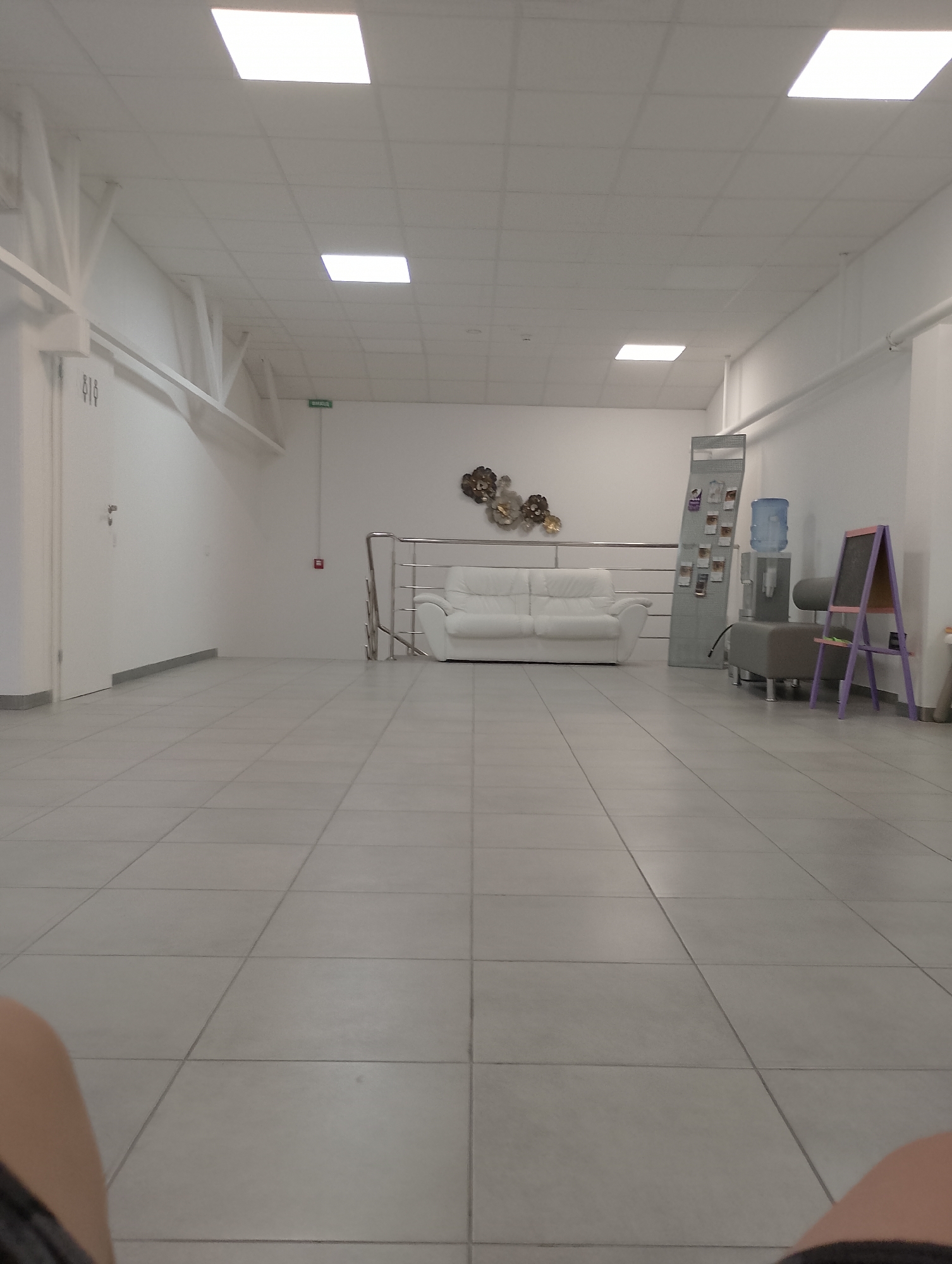 The image depicts an interior hallway or waiting area with a tiled floor and white walls and ceiling. A white sofa is visible in the middle ground, alongside a metal railing and stairs leading up. To the right, there is a water cooler, a brochure display stand, and a purple easel with a blackboard. The overall aesthetic is modern and minimalist. The scene is devoid of any people. The location is identified as Chaiky, Ukraine.