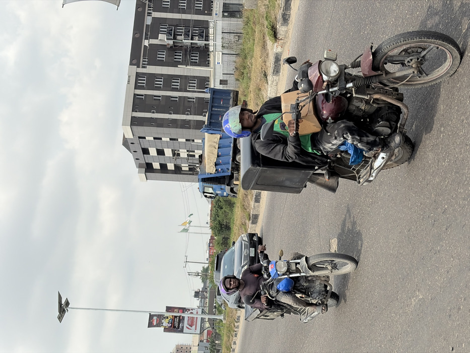 The scene depicts an urban road in Igboefon, Nigeria, under an overcast sky. Two males are operating motorcycles, traveling in the same direction on a paved asphalt road.
In the foreground, a dark-complexioned male is seated on a maroon motorcycle, looking forward. He wears a light blue helmet with a green face shield, a black hooded jacket over a green t-shirt, and dark pants. A large black delivery box is secured to the rear of his motorcycle, and a brown cardboard box rests on the fuel tank area in front of him.
Behind and to the left, another dark-complexioned male is seated on a blue and silver motorcycle, also looking forward. He wears a dark purple helmet, a dark long-sleeved shirt, and light-colored pants. A black, open-top tray structure is mounted on the rear of his vehicle.
A dark-colored sedan is partially visible behind the second motorcycle. Further in the background on the right, a large blue dump truck with a beige tarp covering its bed is present. On the far right, a multi-story building with dark grey/black cladding and white-framed windows stands prominently. To the left, a tall white street light pole equipped with a solar panel array and multiple light fixtures is visible. Two billboards are mounted on another pole behind the first motorcycle, with the top one reading "LUXURY PERFUMES." Two flags, one green and white, the other yellow and white, are visible flying from poles in the mid-distance. Sparse vegetation lines the road shoulders.