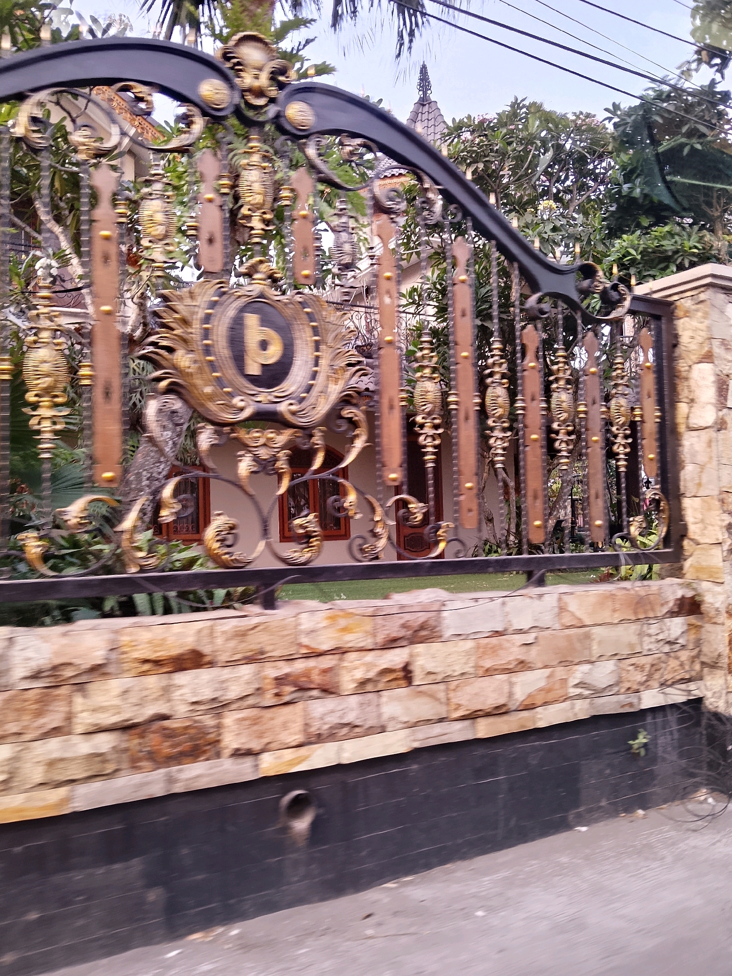 The image depicts a section of an ornate gate in front of a house, likely located in Malang, Indonesia. The gate features intricate black metalwork with gold embellishments, including floral motifs and decorative finials. A circular emblem with a stylized "p" is prominently displayed on the gate. Behind the gate, glimpses of the house's facade can be seen, with wooden-framed windows. Lush green foliage and trees surround the property, with palm fronds visible at the top. The lower portion of the image shows a stone wall and a dark-colored, possibly asphalt, surface in the foreground. The sky is overcast. No people are visible in the scene.