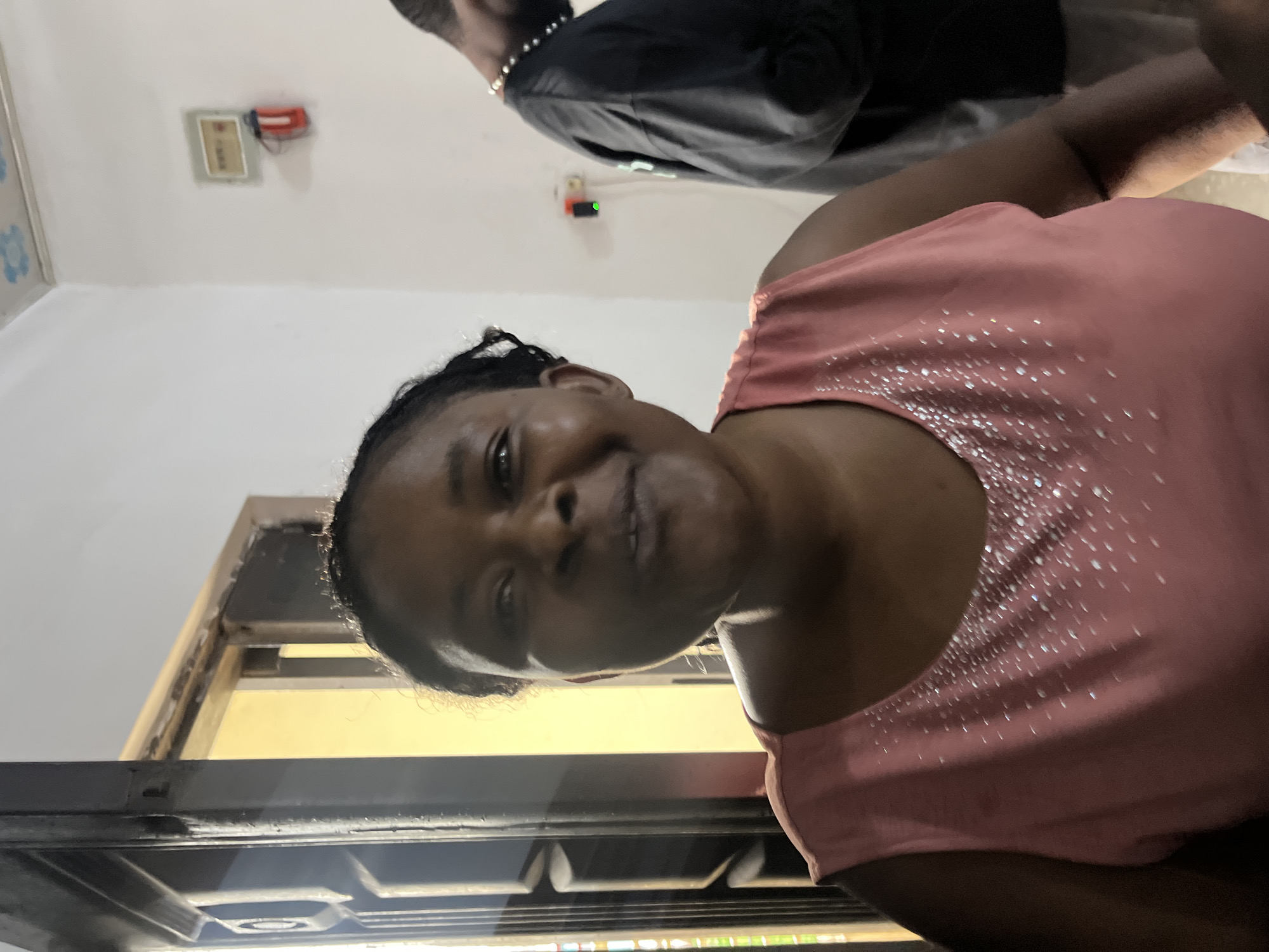 The image shows two people in what appears to be an indoor setting, likely a home or office. One subject is a woman in the foreground, wearing a pink sleeveless top adorned with small, reflective embellishments. She is smiling and looking towards the camera, her head tilted slightly. Her hair is dark and pulled back. The other subject, partially visible in the upper portion of the image, is a person wearing a black t-shirt or jacket, with dark hair and a necklace.
In the background, on a white wall, there is a rectangular object mounted, which appears to be a fire alarm or a similar safety device, with a red component. There is also a window or doorway with a light yellow interior visible through it, suggesting it might be daytime. The overall impression is a casual and friendly interaction, possibly a selfie or a posed photograph. No specific time of day or weather conditions are discernible. The setting appears to be Gura Top, Nigeria.