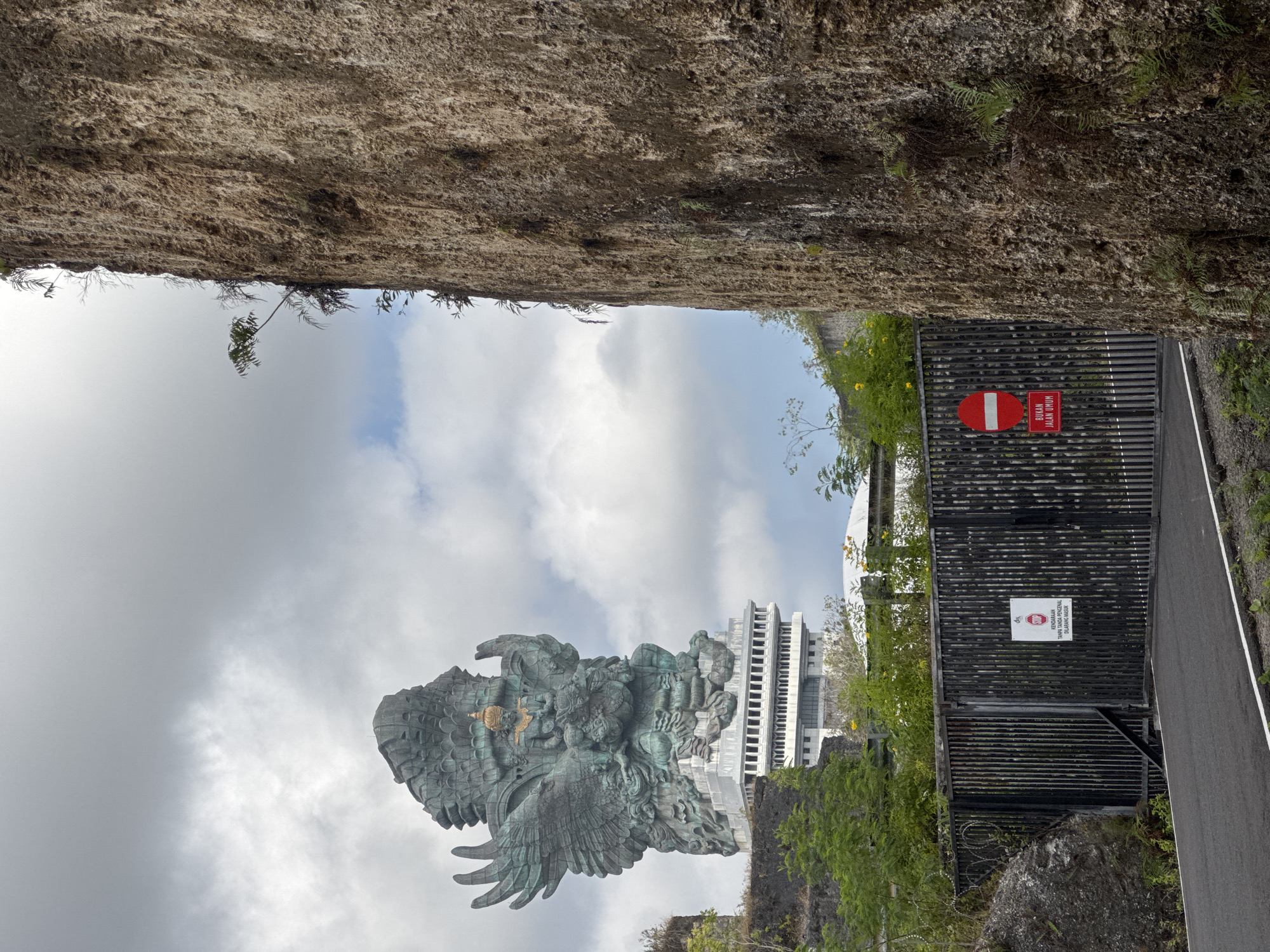 The image shows the Garuda Wisnu Kencana Cultural Park in Ungasan, Kuta Selatan, Indonesia. A massive bronze and copper statue of Garuda, a mythical bird-like creature, is visible in the background against a cloudy sky. The statue depicts Garuda carrying Vishnu. In the foreground, a tall, rough-textured cliff face dominates the right side of the frame. A black metal gate with barbed wire on top is positioned in front of the cliff, partially obscuring the view of the statue. Two signs are attached to the gate; one is a white sign with a red stop symbol and text in Indonesian, and the other is a red rectangular sign with a white horizontal bar and Indonesian text below it. A paved road leads up to the gate, and various green plants and trees are scattered around the scene, growing both in front of and on the cliff face.