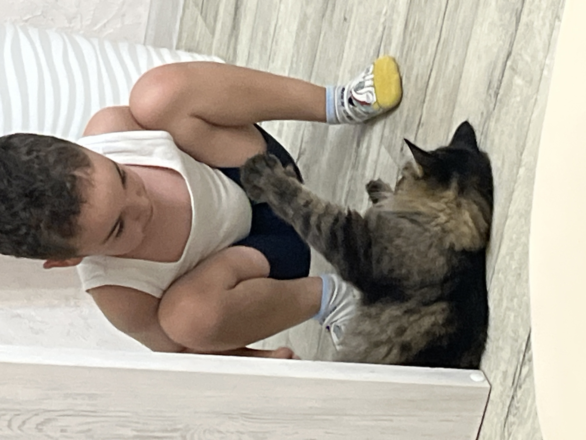 A young child in a white tank top and dark shorts is crouching on a wooden floor next to a fluffy tabby cat. The child's face is obscured as they look down at the cat, which is lying on its back with its paws in the air. The child is wearing socks with a yellow toe and a white and blue pattern. A white wooden structure is on the left side of the frame. The background consists of a light-colored wall with a wavy striped pattern. The scene is indoors, possibly in a residential setting in Krasnyye Barrikady, Russia.