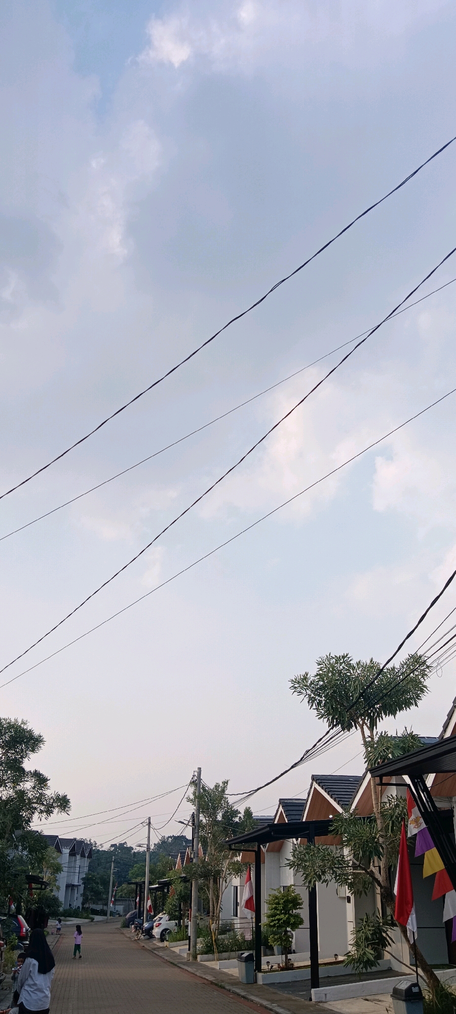 A low-angle, vertical shot captures a street scene in Kemang, Indonesia. The sky, filled with pale, wispy clouds, dominates the upper portion of the image, crisscrossed by several dark electrical wires. Below the sky, a row of modern houses with dark tiled roofs and white walls lines the street. Some houses feature awnings supported by black poles, and small trees and plants are visible in front of them. Several small Indonesian flags, red and white, are hung on poles or attached to the awnings. A few cars are parked on the side of the paved street, and some people, including two women and a child, are walking. The overall impression is one of a quiet residential street under a slightly overcast sky.