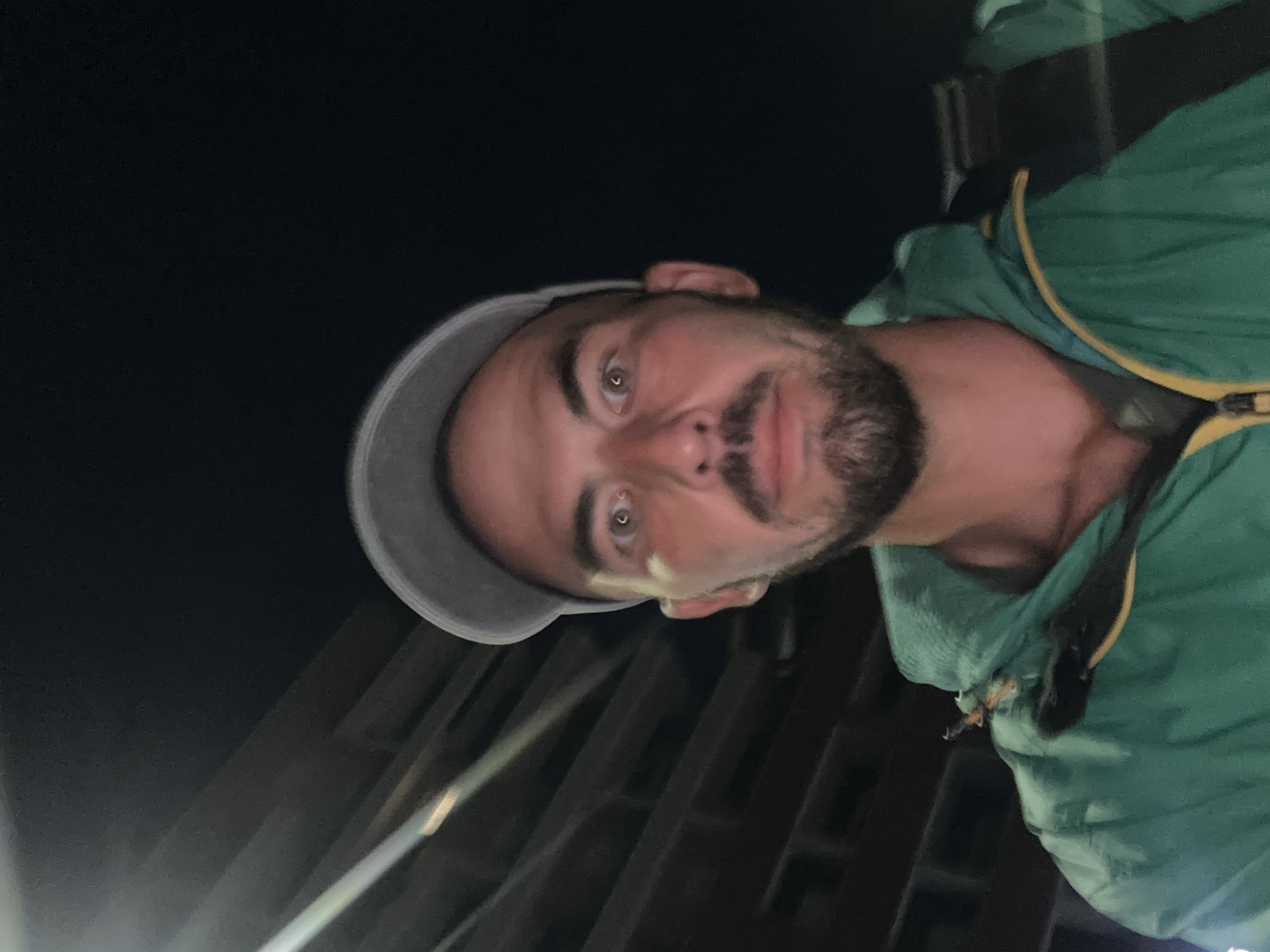 A man wearing a green jacket and a gray baseball cap is looking at the camera at night. The background shows a dark, possibly brick building with horizontal lines. The man has a beard and open eyes. A black backpack strap is visible over his right shoulder.