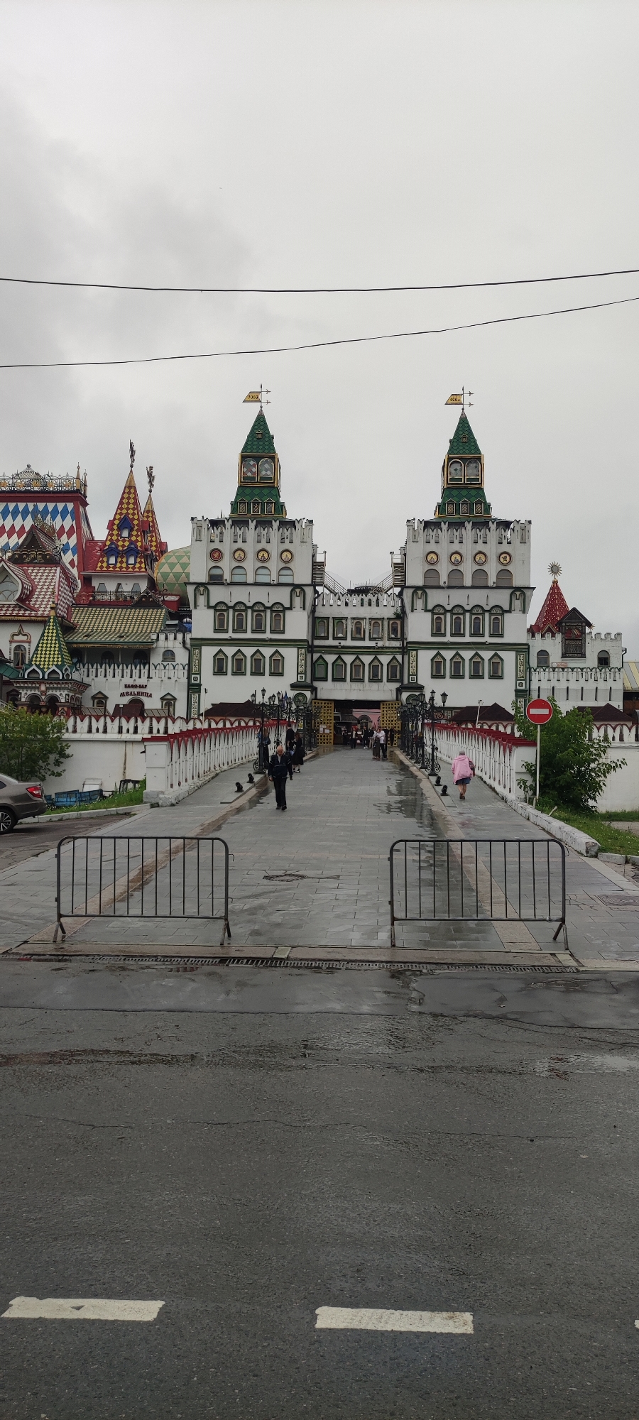 The image captures the Izmaylovo Kremlin, a vibrant, fairytale-like complex located in Moscow, Russia. The Kremlin's distinctive architecture, characterized by colorful towers, intricate rooflines, and decorative facades, is the central focus. Two prominent towers, adorned with green roofs and topped with weather vanes indicating the year "2013", stand symmetrically. A wide, paved walkway, wet from recent rain, leads towards the entrance of the complex. Temporary metal barriers are placed on the walkway in the foreground, suggesting a controlled access or event. Several people are visible walking across the bridge and towards the Kremlin, including a man in a dark jacket and a person wearing a bright pink backpack. A car is parked on the left side of the image, and a red and white striped fence lines the walkway. The sky is overcast, contributing to the muted lighting of the scene.