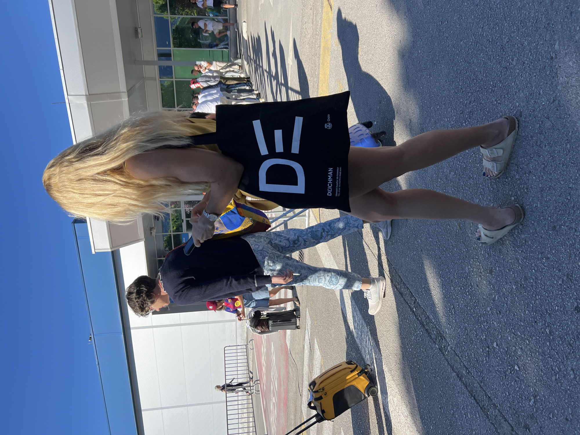 The image captures a sunny outdoor scene, likely an airport, with a focus on individuals and their luggage. A woman with long blonde hair, wearing a yellow top and light-colored sandals, is in the foreground, holding a black tote bag with the letters "DE" prominently displayed. The bag also features text that reads "DEICHMAN Oslo" and a tagline in Norwegian. She is looking down at her phone, which she is holding in her left hand.
To her left, a person wearing a dark navy sweater and patterned blue pants, along with white sneakers, is also looking towards their phone. This individual has glasses and short, dark hair. They are carrying a cigarette in their right hand. In front of this person, a yellow suitcase with black wheels is positioned on the asphalt.
In the background, several other people are visible, some with luggage. A man in a white shirt and shorts is standing in line. Another person is seen walking away from the camera towards a metal fence. The environment appears to be a paved area with shadows cast by the sun, suggesting it is a bright day. The architecture in the background features a modern building with large windows. The scene suggests a moment of travel or transit, with people engaged in personal activities while waiting or moving through the area. The location is identified as Mrčevac, Montenegro.
