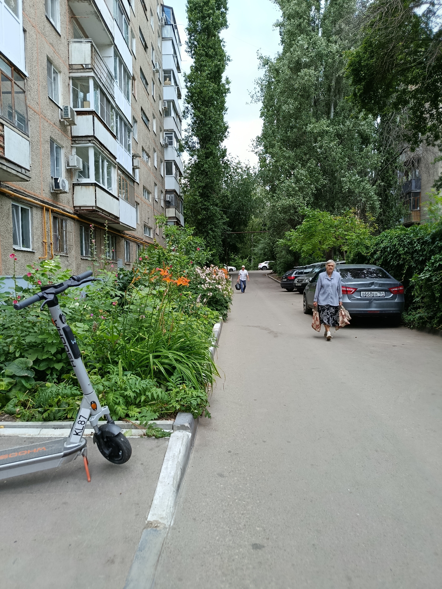 An eye-level, medium shot captures an outdoor scene likely in Saratov, Russia, featuring a residential building, a pathway, parked cars, and vegetation. In the foreground to the left, a grey electric scooter stands on the asphalt pathway. The scooter has a black handlebar, orange accents, and "KL87" marked on its front fork. The pathway curves towards the background, leading to a parking area where several cars are parked. A woman in a blue cardigan and a patterned skirt is walking away from the viewer, carrying two grocery bags. Further in the background, another person, possibly a man wearing a white shirt and dark trousers, is walking in the opposite direction, carrying a blue bag. The residential building on the left is multi-story with several balconies, and a row of green plants and orange flowers runs along its base. Tall trees and dense green foliage line the right side of the pathway and the parking area. The sky is overcast.