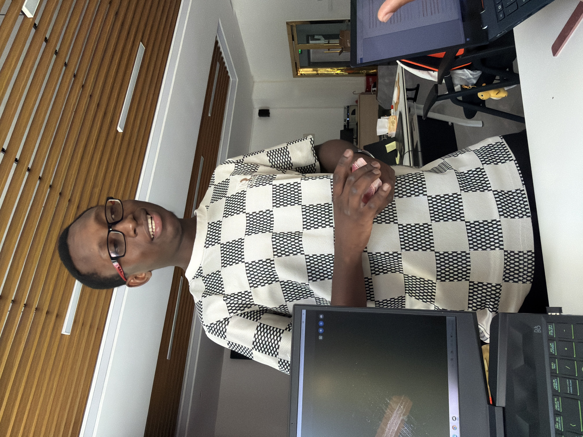 A young Black male wearing glasses and a checkered shirt stands in an office setting. He is holding some pink items in his hands. Two laptops are visible on a desk in front of him, and a yellow duck keychain hangs from a chair in the background. The office has a modern design with wood-paneled ceilings and fluorescent lighting. The provided context places the image in Jiangjin District, Chongqing, China.