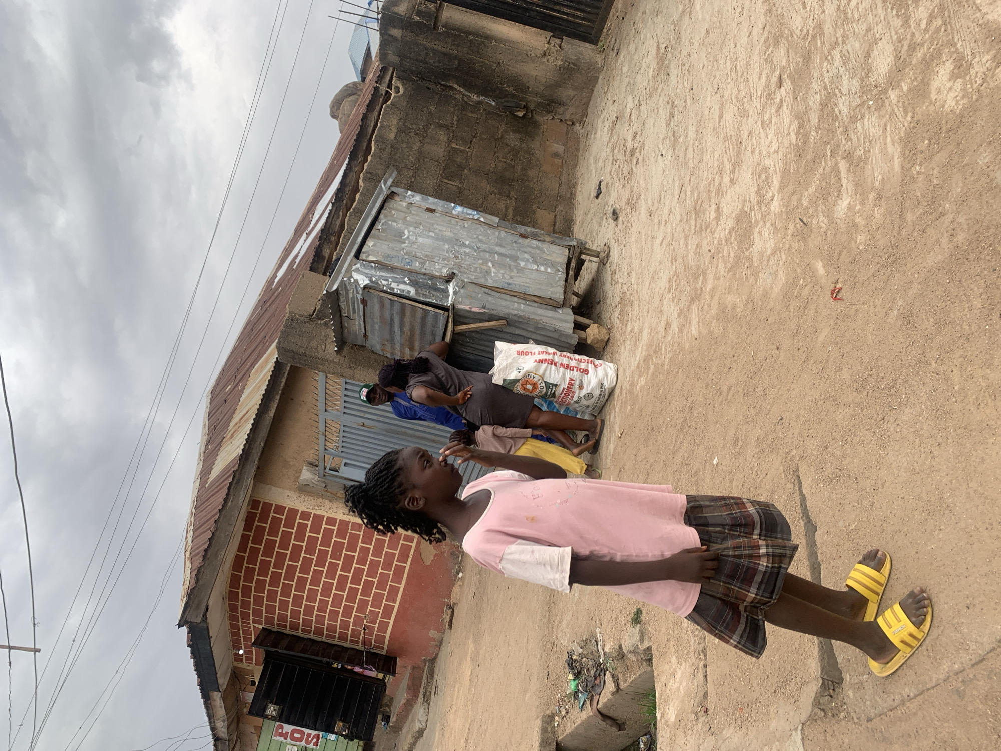 A young girl with braided hair is standing on a concrete walkway next to a dirt road in what appears to be a residential area. She is wearing a light pink shirt, a plaid skirt, and yellow sandals. Behind her, two adults are standing next to a building with a corrugated metal extension and a large white sack labeled "GOLDEN PENNY ENRICHED WHEAT FLOUR". The building has a brick facade. The sky is overcast. Electrical lines are visible overhead. There are various items discarded on the side of the road near a drainage ditch. The scene is consistent with a residential area in Jos, Nigeria.