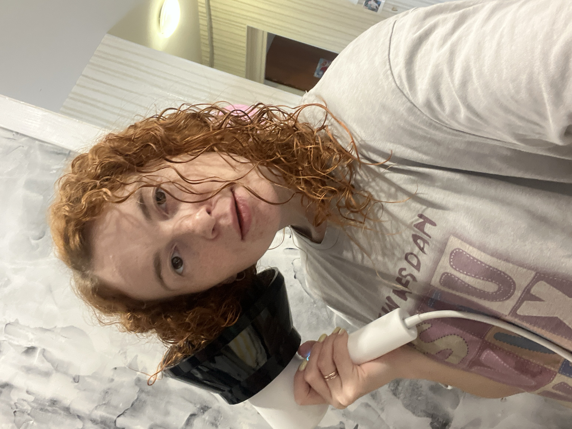 A woman with reddish-brown curly hair is holding a white hair dryer with a black diffuser attachment to her head. She is looking at the camera with a neutral expression. She is wearing a light gray t-shirt with a graphic design. She has a gold ring on her finger and yellow nail polish.
The background appears to be a bathroom. There is a marble-like wall pattern visible. The location in Chaikovsky, Russia is not discernible from the image.