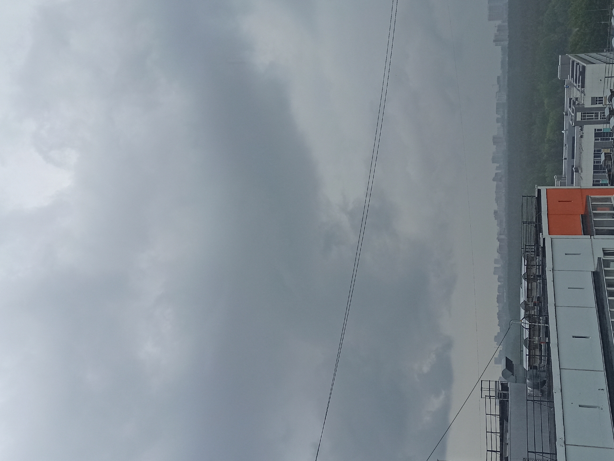 The image shows a cityscape under a cloudy sky. A tall building is visible on the right side of the frame, with an orange section on its top. Power lines run diagonally across the sky. In the distance, there's a view of other buildings partially obscured by the weather conditions.
