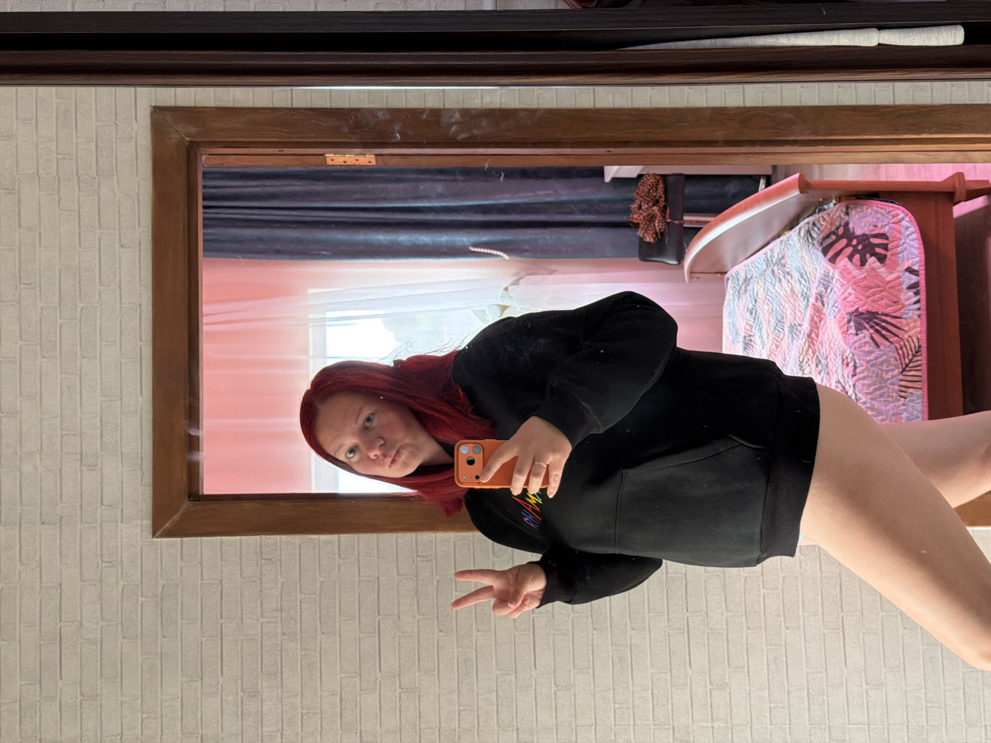 A photograph depicts a person with red hair, likely female, taking a selfie in front of a mirror. The person is wearing a black hoodie and is making a peace sign with their hand. The mirror's reflection shows a bed with a patterned blanket and what appears to be a headboard, suggesting a bedroom setting. The walls around the mirror are brick patterned, and a wooden frame surrounds the mirror itself. The location is not specified or discernible from the image.