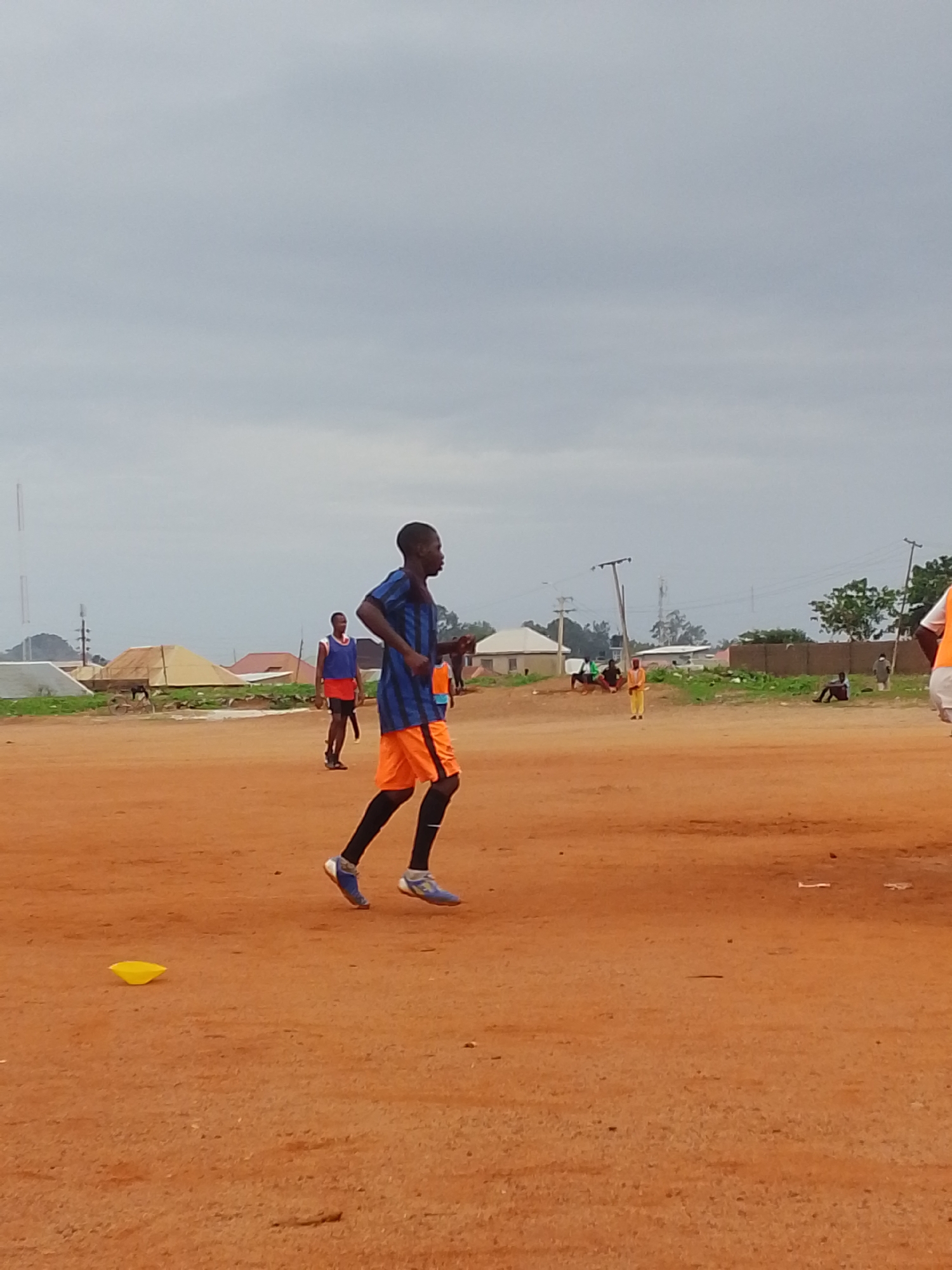 On a cloudy day in Bukuru, Nigeria, a young man wearing a blue and black striped jersey, orange shorts, black socks, and blue sneakers runs across a dirt field. Another individual in a red and blue vest with dark shorts stands in the background. There are several people sitting or standing on the periphery of the field. A yellow plastic cone sits on the ground. In the background, there are houses, a pile of sand, power lines, and a radio tower.