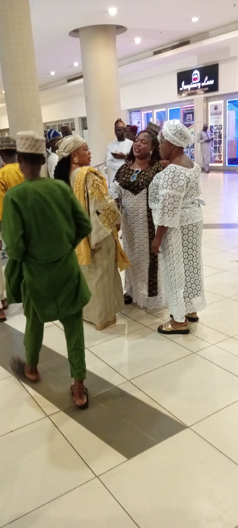 In Gandu Albasa, Nigeria, a group of individuals are gathered indoors, possibly at a shopping mall given the tiled floor and overhead lighting. Three women in the foreground are engaged in conversation. The woman on the far left wears a light-colored headscarf and a gold and cream patterned outfit. Next to her, a woman in a white lace dress with a black and white patterned neckline is speaking animatedly. To her right, another woman in a white lace dress and matching headscarf also appears to be participating in the discussion. A man in a green traditional outfit is walking away from the women, facing towards the left of the frame. In the background, other individuals in traditional attire can be seen, and a sign for "Imaginary Lane LED Virtual Reality Ride" is visible on the wall. The environment suggests a public indoor space with commercial establishments.