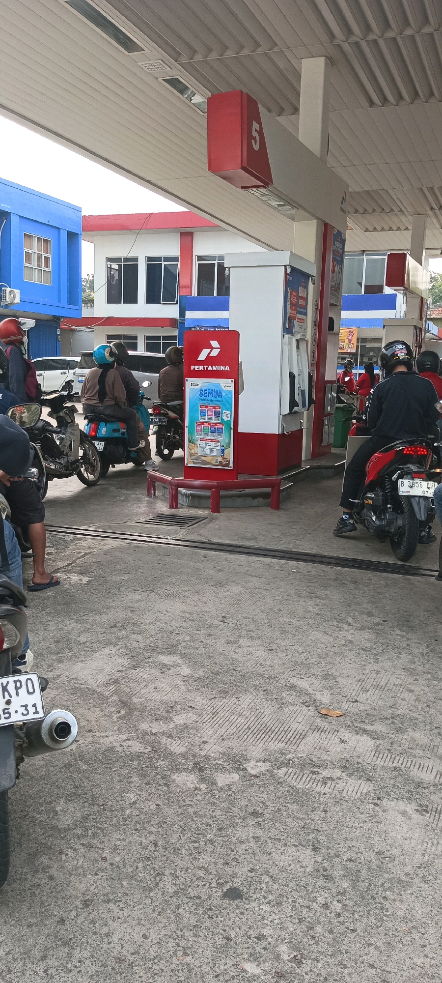 The image shows a Pertamina gas station in East Jakarta, Indonesia. Several motorcycles and their riders are visible at the pumps, suggesting they are refueling. The scene is outdoors, with a covered canopy over the gas pumps. In the background, a blue building with windows and a red and white building are also present. The ground is paved concrete with visible tire marks and some debris. The overall atmosphere appears to be a busy, everyday scene at a public refueling station.
