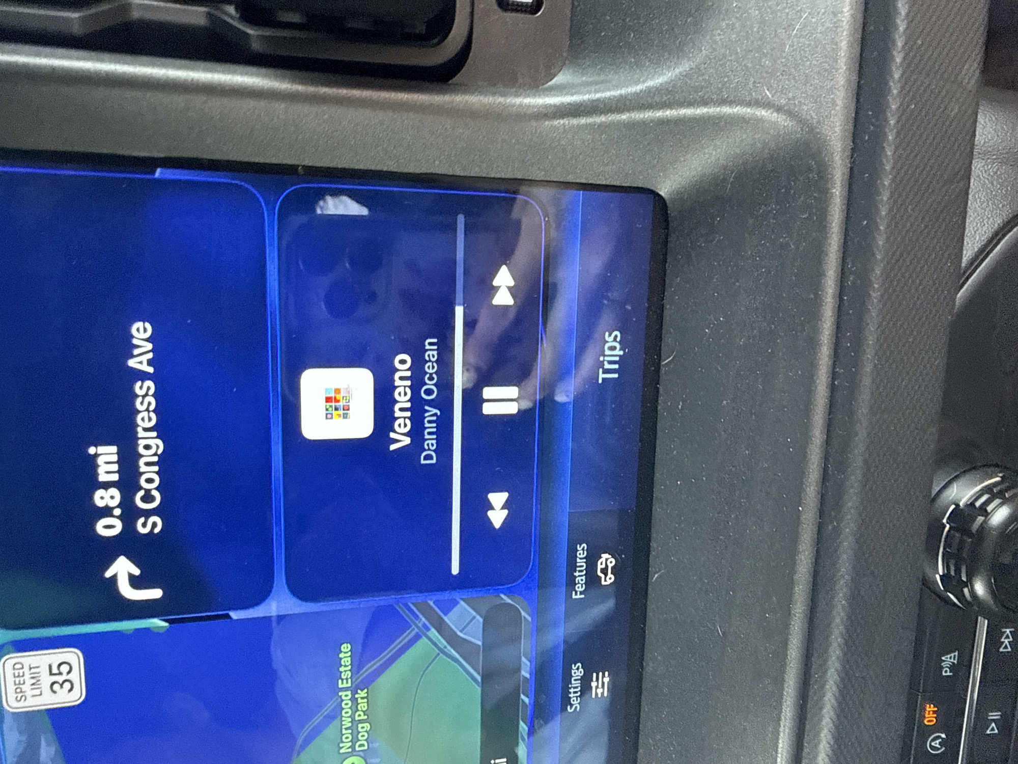 The image depicts a car's infotainment screen. The screen shows navigation with a current location 0.8 miles from S Congress Avenue. There is a speed limit sign stating 35 miles per hour. Also displayed is a music app playing a song named "Veneno" by Danny Ocean. The screen also shows other menu options such as settings, features, and trips.