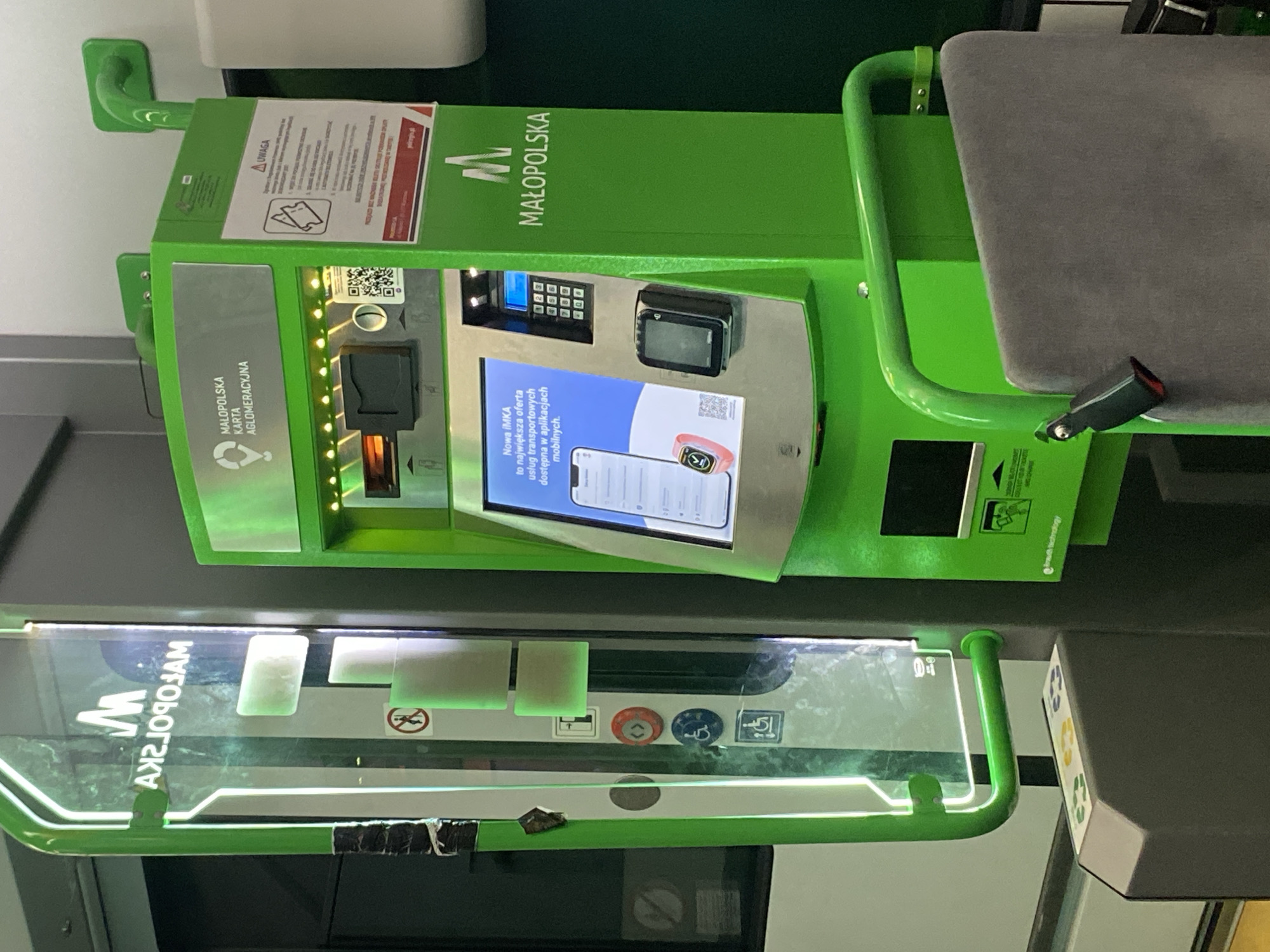 In Kępa Bogumiłowicka, Poland, a bright green ticket vending machine stands prominently. The machine features a touchscreen display with information in Polish about the Małopolska region and its public transport services. Below the screen, a keypad and a card reader are visible, suggesting options for ticket purchase and payment. To the left of the ticket machine, a glass panel displays a green "M" logo and the words "MAŁOPOLSKA KARTA AGLOMERACYJNA" in reverse, indicating it's part of the Małopolska Agglomeration Card system. Various symbols, including a "no smoking" sign and accessibility icons, are present on the panel. The machine is situated in an indoor environment, possibly a train station or public transport hub, with hints of other structures and lighting in the background. A grey upholstered seat is attached to the side of the machine.