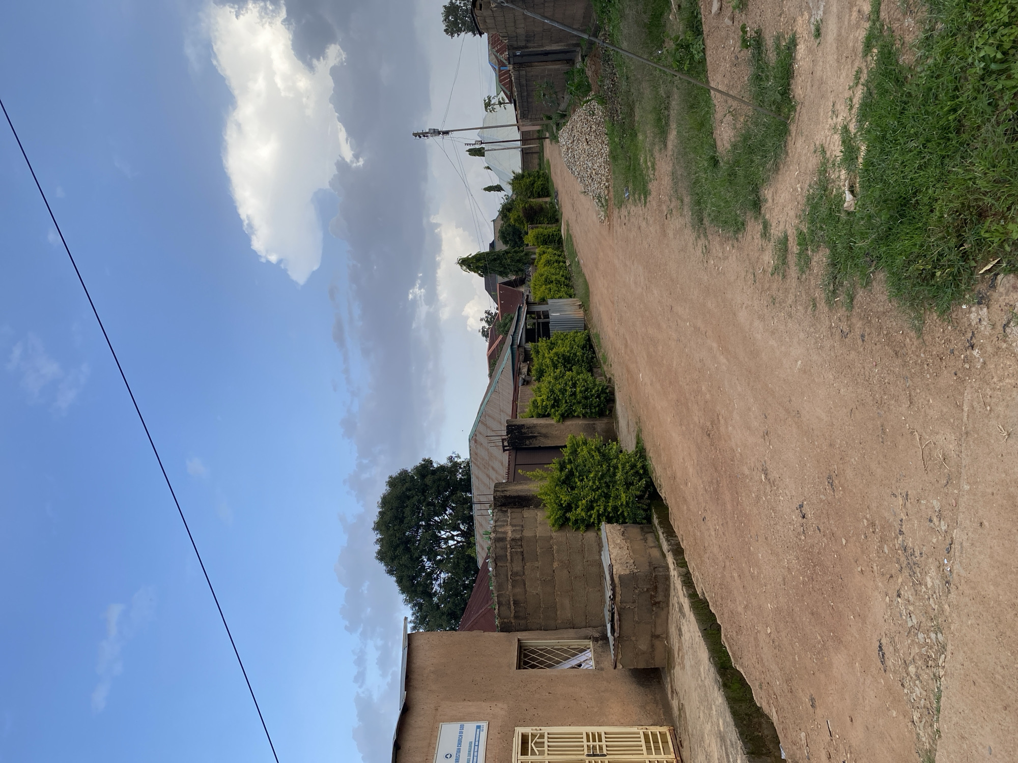 The image depicts a dirt road in Gura Top, Nigeria, lined with houses and vegetation. On the left, a building with a sign reading "Christian Church of God" is visible, featuring a light yellow gate and a window. Adjacent to it is a low brick structure. The road curves to the right, flanked by hedges and fences. Several houses with corrugated metal roofs are scattered along the road, some with visible windows and doors. A pile of light-colored stones sits on the right side of the road. In the background, trees and utility poles with wires are present. The sky is a mix of blue with scattered white and gray clouds. The ground is dusty, with patches of green grass growing along the sides of the road and in some areas of the foreground.