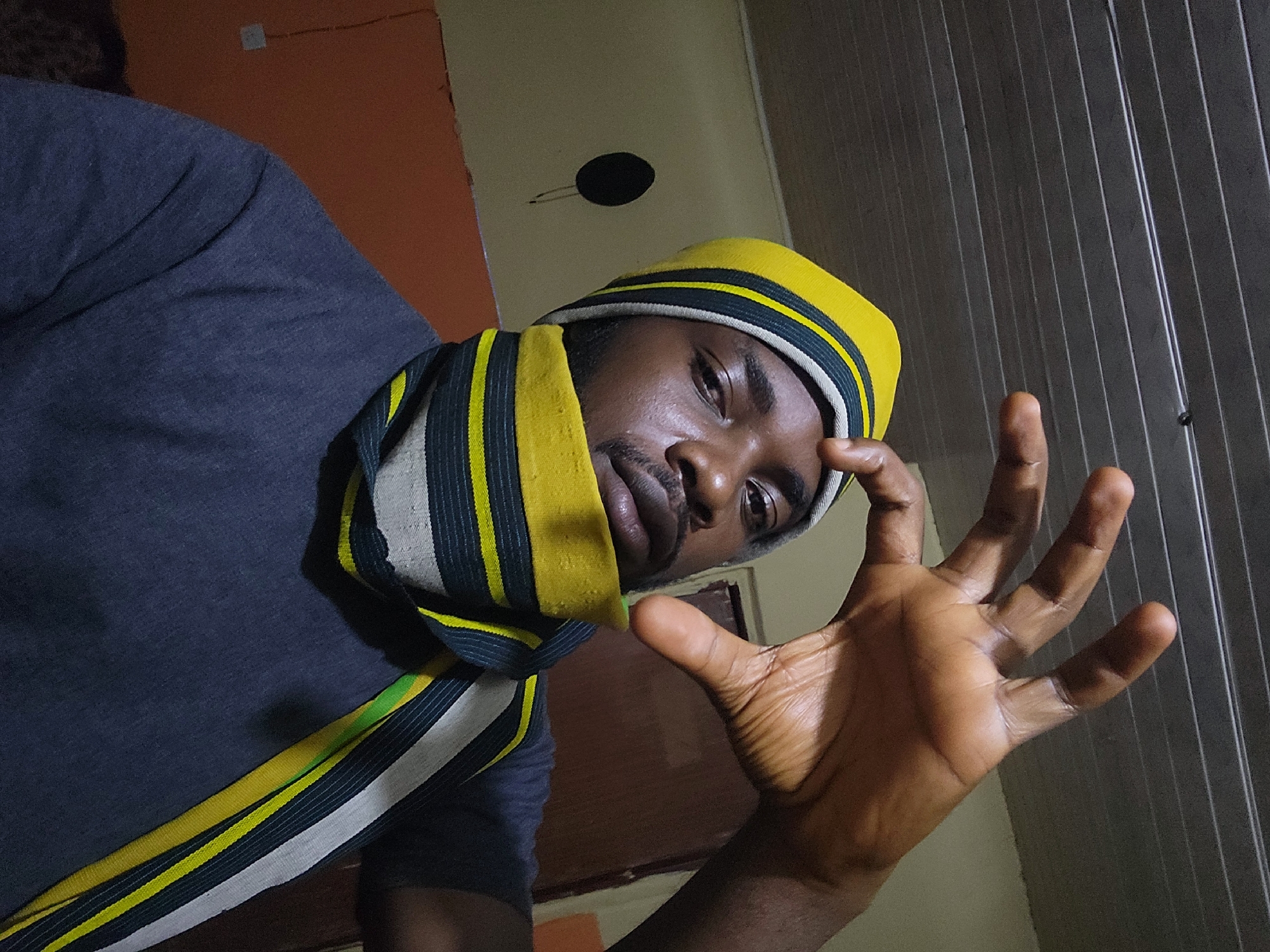 The image depicts a close-up, selfie-style shot of a Black male wearing a yellow and black striped beanie and a matching scarf. His right hand is raised towards the camera, with fingers spread. He has a beard and is looking directly at the camera. The background appears to be an indoor setting, with a textured ceiling and a wall painted in an orangish hue. A dark, circular object is visible on the wall to the left. The overall lighting suggests an indoor environment, possibly at night or in a dimly lit room. There is no indication of the specific location within Jos, Nigeria, beyond it being an indoor residential or personal space.