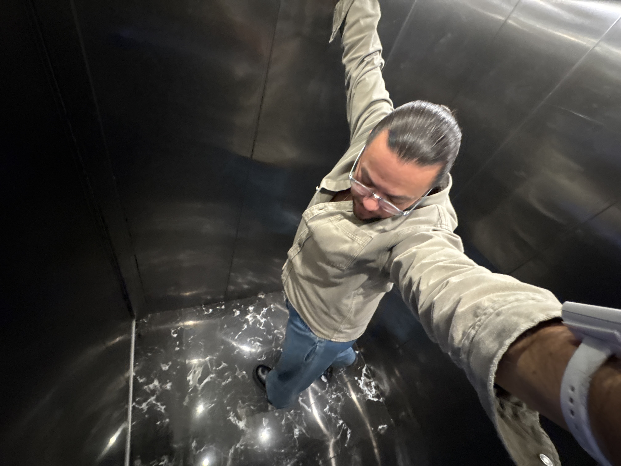 A man is taking a selfie from a low angle in what appears to be an elevator. The elevator walls are shiny, dark metal and the floor is black and white marble. The man has dark hair slicked back, glasses, and is wearing a light grey jacket and blue jeans. He is wearing a white bracelet on his wrist. The location is unknown and cannot be determined from the image.