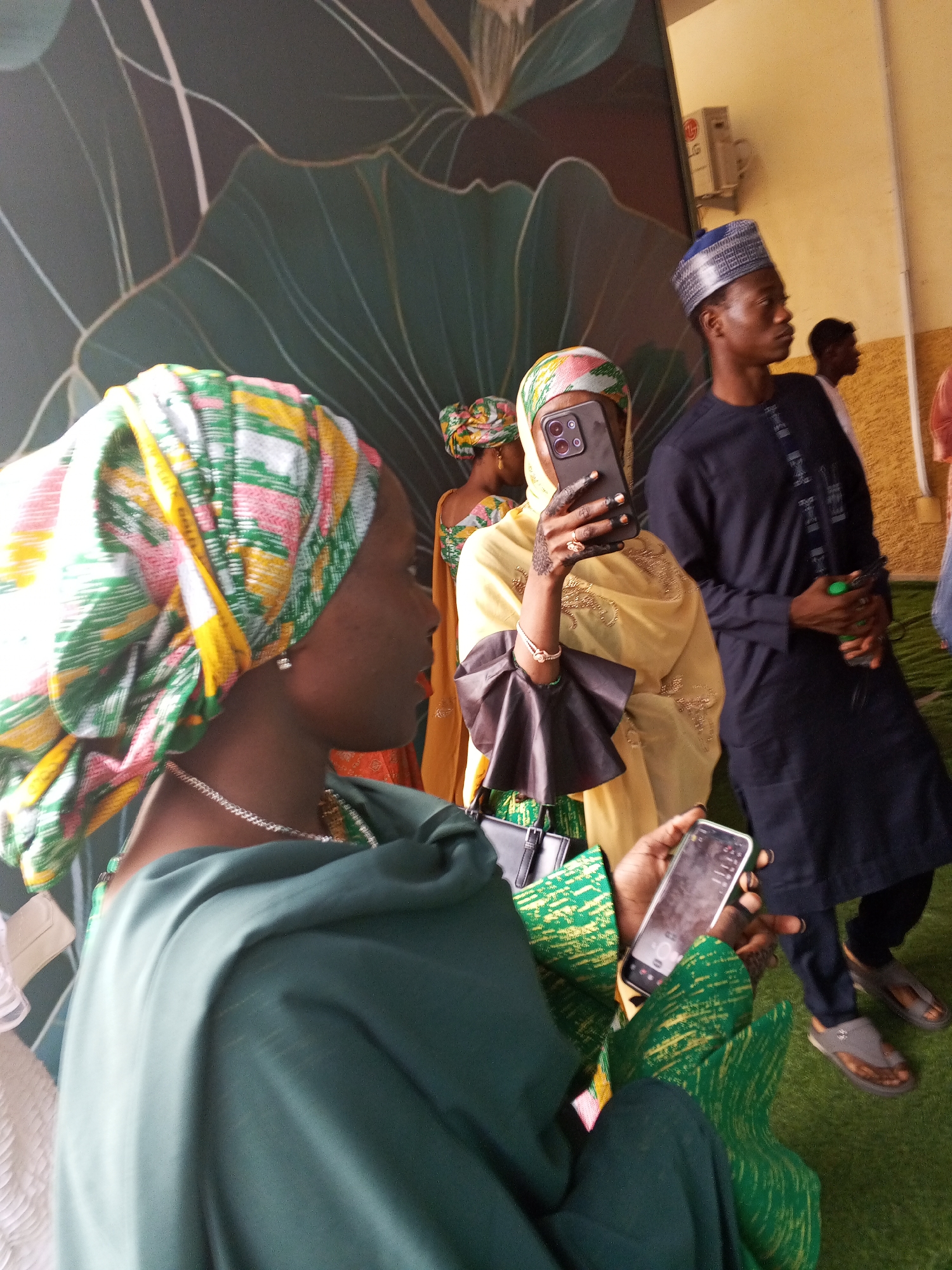 In Abuja, Nigeria, a group of people are gathered, possibly at an outdoor event or public space with a mural depicting large, abstract floral shapes on a dark background. In the foreground, a woman wearing a colorful headscarf and a green garment is holding a smartphone, seemingly capturing a photo or video. To her right, another woman in a yellow outfit and a patterned headscarf is also holding a smartphone. Behind them, a man in a dark blue traditional outfit and a blue cap is visible, looking to the right. Another man in the background is partially obscured. The ground appears to be covered in a green, textured material, possibly artificial turf. The overall atmosphere suggests a social gathering, possibly a celebration or a public event.
