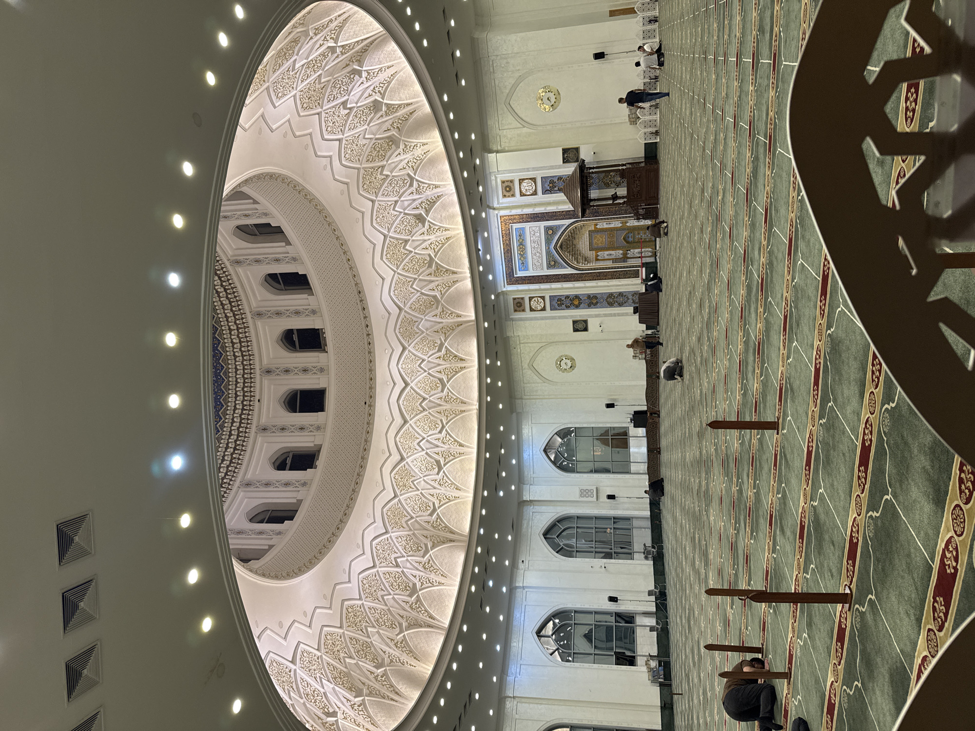 The image depicts the interior of a mosque, likely the Minor Mosque in Tashkent, Uzbekistan, distinguished by its white marble facade and intricate Islamic decorations. The mosque features a large dome with ornate geometric patterns, illuminated by numerous lights. The prayer hall is covered with a green carpet marked with rows, likely indicating individual prayer spaces. Several individuals are present, some kneeling in prayer, while others stand or sit. A wooden minbar (pulpit) is visible at the front of the hall. The architecture includes arched windows and decorative panels along the walls.
