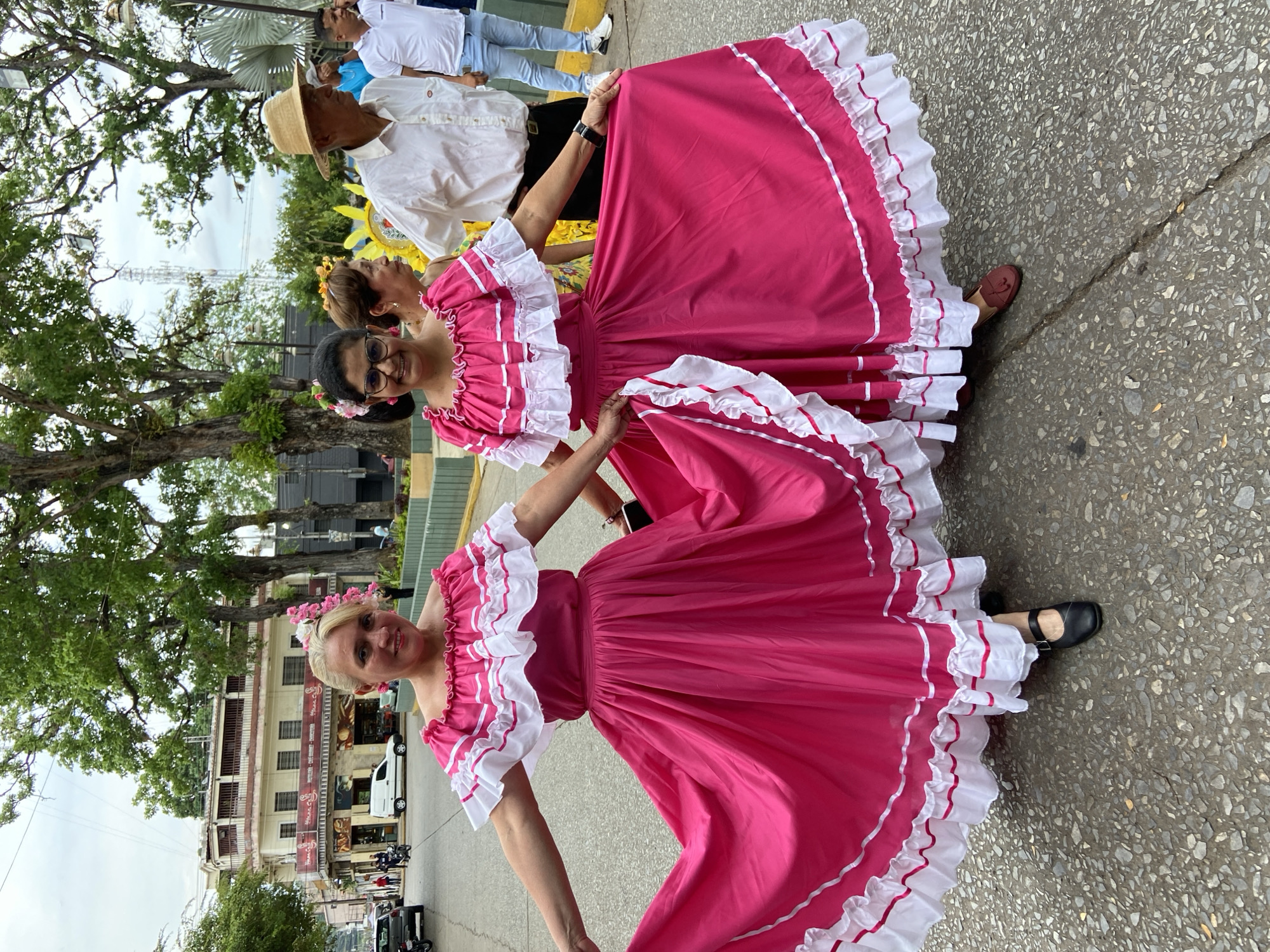 Three women in pink dresses with white ruffles stand on a street in Tinaquillo, Venezuela. Two of the women have flower crowns. Cars and motorcycles are parked on the street in the background, and a building with multiple stories is also visible. Other people are walking nearby.