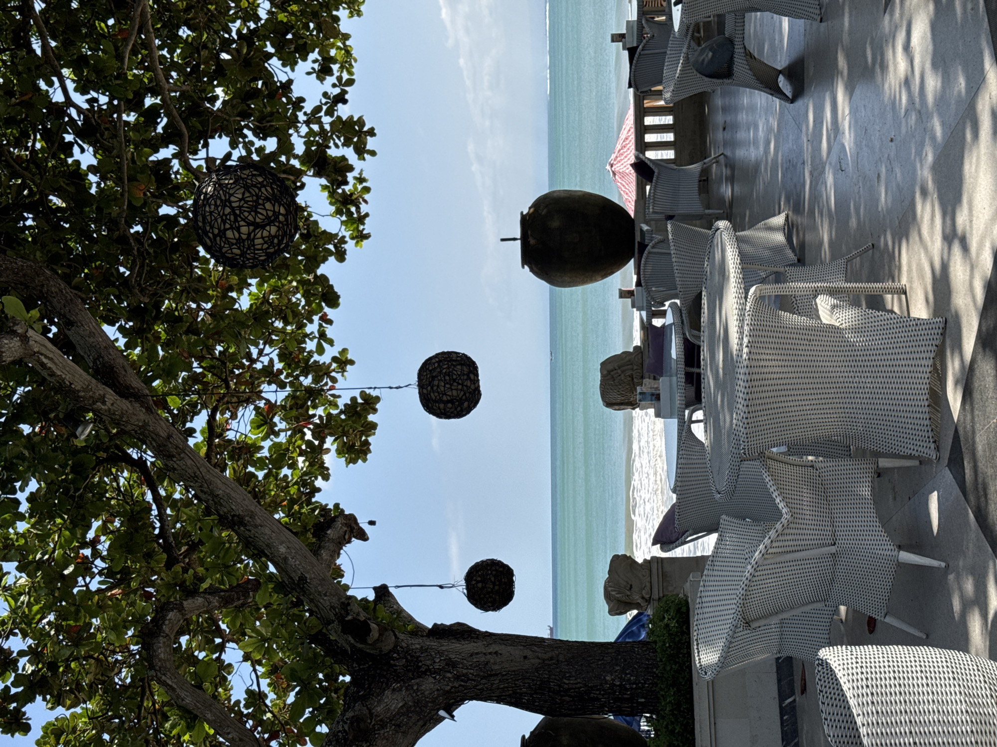 The image shows an outdoor setting in Kuta Selatan, Indonesia, featuring a partially visible tree with hanging decorative spherical lights, a view of the ocean, and outdoor seating on a patio. The tree is to the left, with green leaves and dark branches against a light blue sky. Three round, woven lights hang from the tree's branches. The ocean stretches across the mid-ground with turquoise water under a bright sky. To the right, there's a patio area with white wicker chairs and tables, suggesting a restaurant or café setting. A large earthen jar sits on the patio near the seating, acting as a decorative element. The scene is brightly lit, indicating daylight. The overall setting evokes a relaxed, beachfront ambiance.