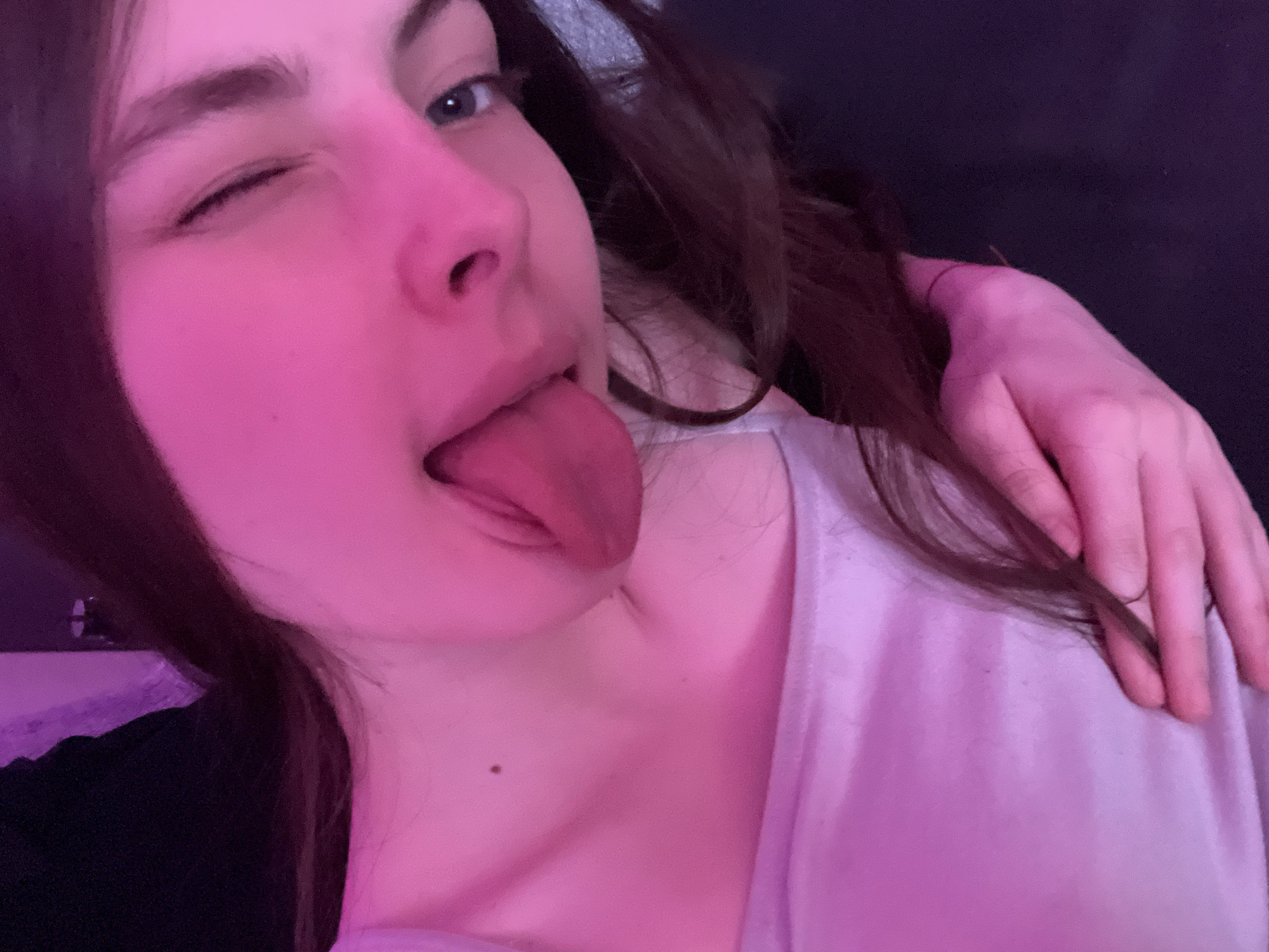 A young woman is positioned close to the camera, possibly in a selfie. Her brown hair is loose, and she is winking with her left eye while sticking her tongue out. The lighting is dominated by a pink hue. She is wearing a white shirt, and another hand is resting on her shoulder. The background is dark and indistinct. There is no information to suggest this photo was taken in Saint Petersburg, Russia.